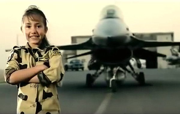 الصورة تظهر فتاة صغيرة تبتسم وتقف بفخر أمام طائرة مقاتلة. ترتدي زيًا عسكريًا يشبه الزي الذي يرتديه الجنود، ويبدو أنها تظهر الثقة والاعتزاز. خلفها، توجد الطائرة، مما يعطي انطباعًا عن القوة والجرأة. الأجواء في الصورة تبدو عسكرية، مع خلفية ترابية وألوان تدل على بيئة القاعدة العسكرية.