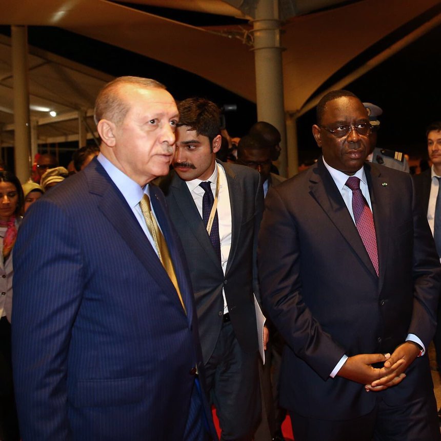 The image shows two prominent figures walking together in a formal setting. One is wearing a dark suit with a light blue striped tie, and the other is dressed in a dark suit with a maroon tie. They appear to be engaged in conversation or deep in thought. In the background, there is a ceremonial guard dressed in a bright red uniform, standing at attention, adding a sense of formality to the scene. The environment looks like an official venue, possibly an airport or a conference center, illuminated by soft lighting.