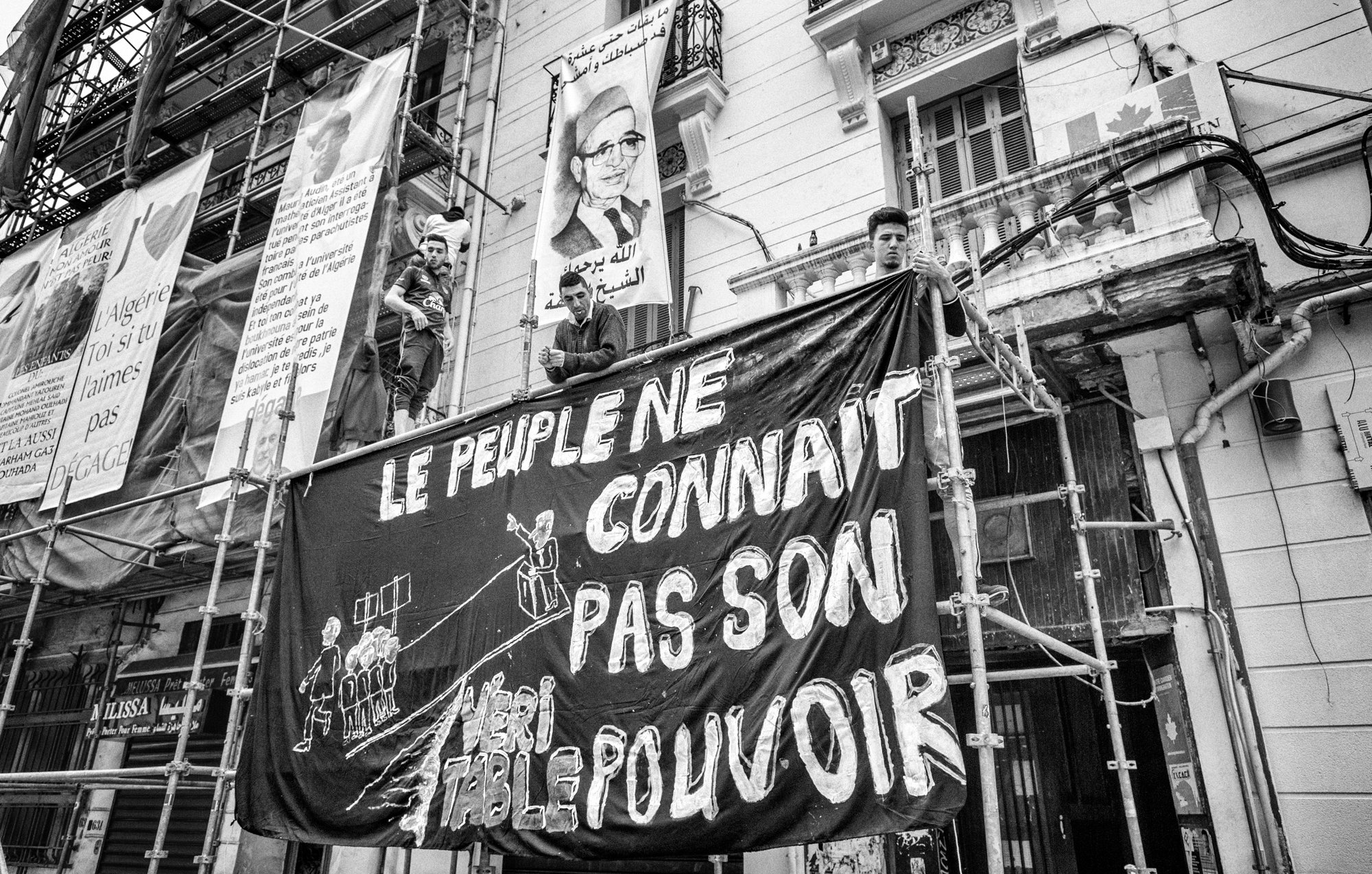 L'image présente une scène en noir et blanc où des personnes sont perchées sur un échafaudage face à un bâtiment. Ils tiennent une grande bannière sur laquelle est écrit en lettres blanches : "LE PEUPLE NE CONNAÎT PAS SON POUVOIR". En arrière-plan, on peut voir des affiches et des motifs typiques d'un environnement urbain. Cette composition évoque un sentiment de protestation ou de revendication sociale.