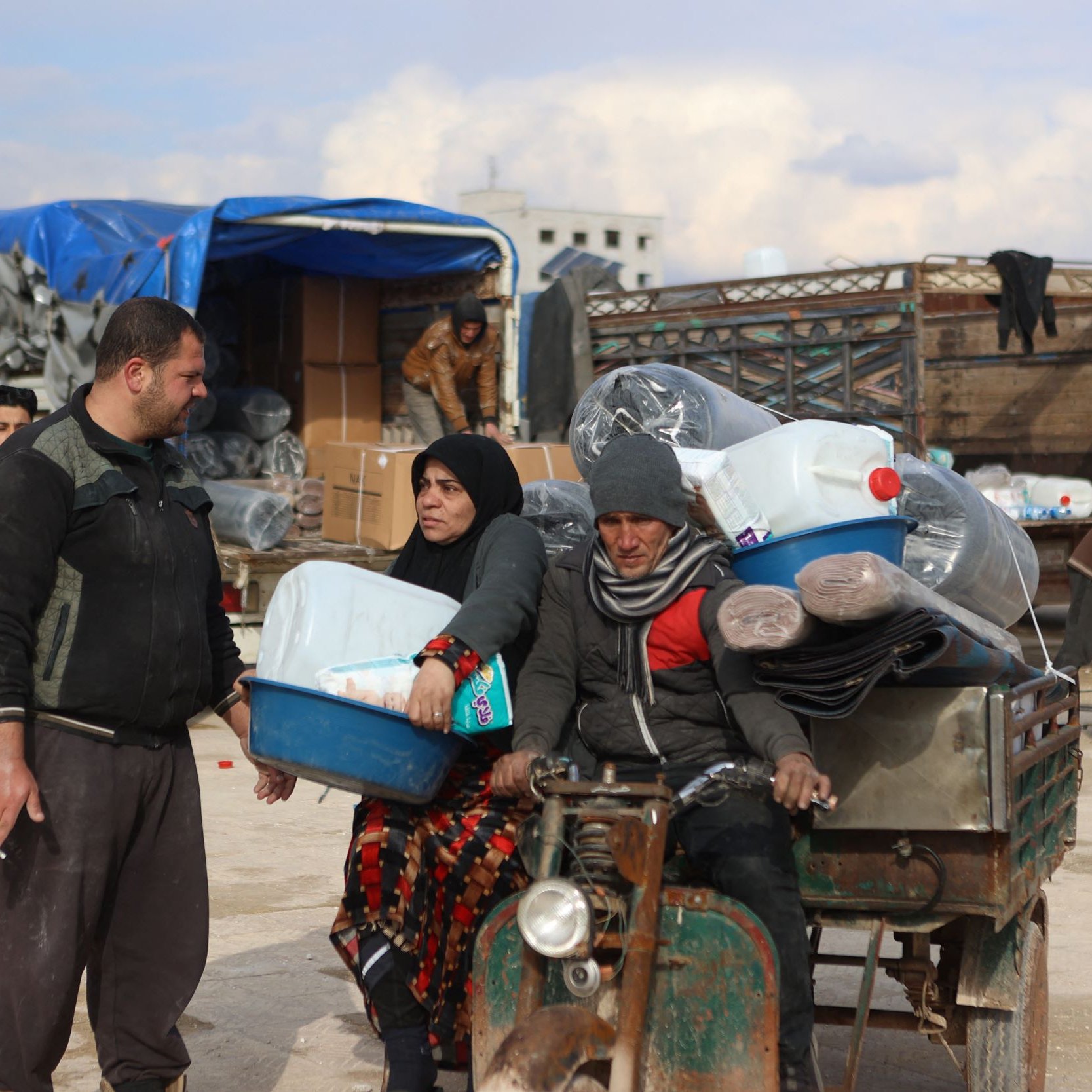 L'image montre une scène de marché ou de distribution dans un environnement rural. On y voit un homme et une femme discutant, tandis qu'un autre homme est assis sur un véhicule à trois roues, probablement en train de transporter des marchandises. Le décor entoure des containers en plastique, et plusieurs personnes s'affairent à décharger ou à déplacer des articles. Le ciel est nuageux, suggérant une atmosphère d'activité et d'échange.