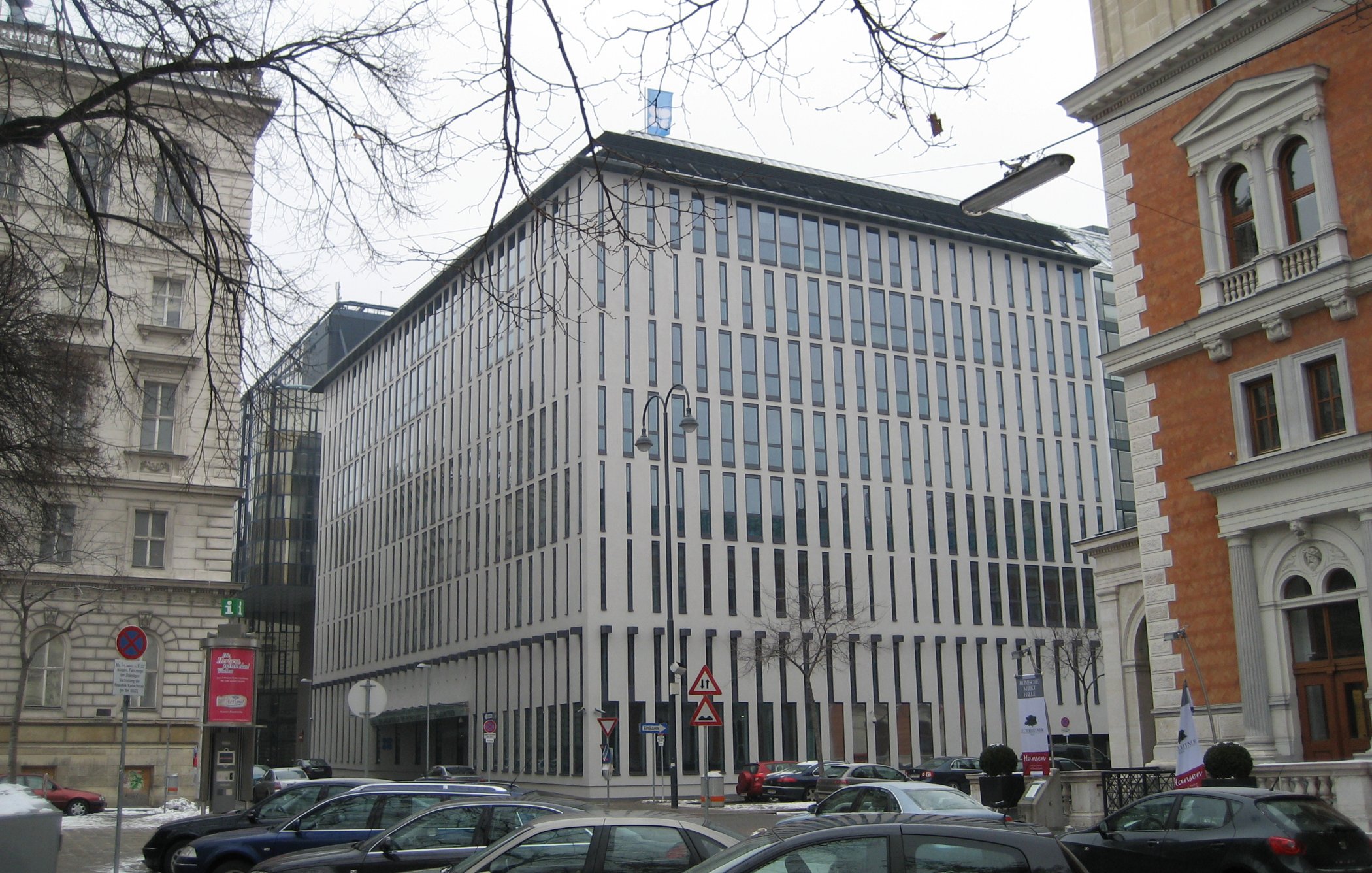 L'image montre un bâtiment moderne avec une façade composée de lignes verticales blanches. Il se trouve à proximité d'autres structures architecturales, dont l'une semble plus ancienne, en pierre avec des ornements. Le paysage est urbain et il y a plusieurs voitures garées au premier plan. Le sol est couvert de neige, ce qui suggère une ambiance hivernale. Le ciel est nuageux, ajoutant à la scène une atmosphère sobre.