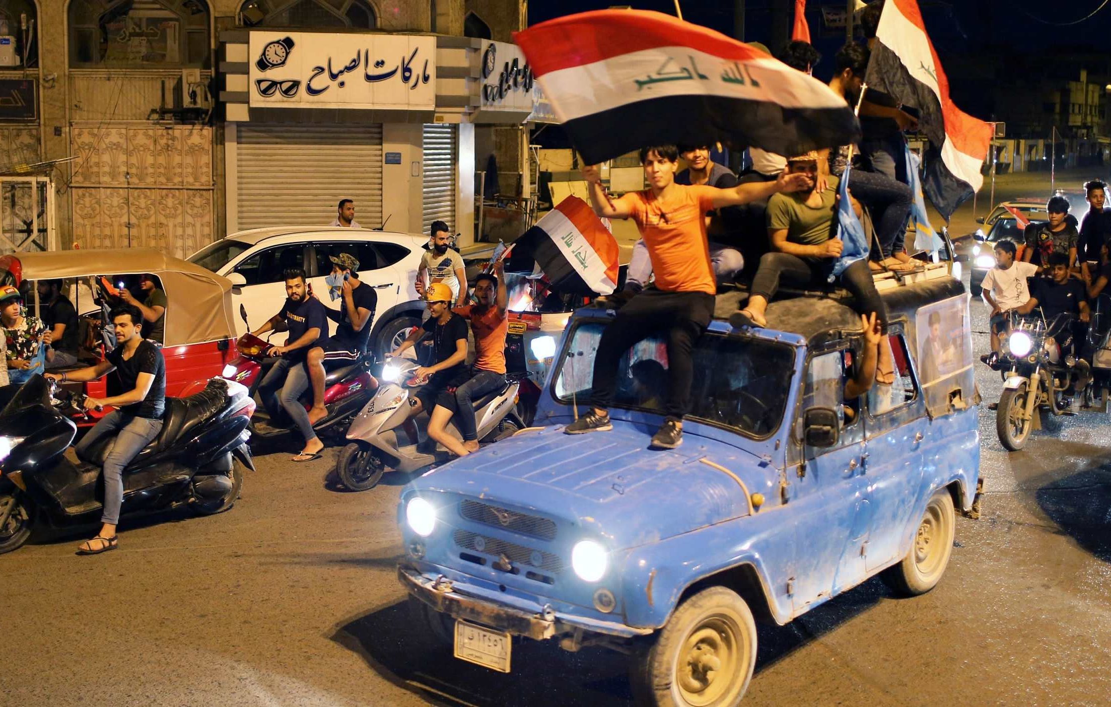 در این تصویر، گروهی از جوانان در یک خیابان در حال شادی و جشن هستند. آنها بر روی یک خودروِ آبی رنگ نشسته‌اند و پرچم عراق را در دست دارند. تعدادی موتورسیکلت نیز در کنار خودرو در حال حرکت هستند. جوانان با شور و هیجان به جشن و شادی پرداخته و فضای پرانرژی و شادابی را ایجاد کرده‌اند. در پس‌زمینه، ساختمان‌ها و فروشگاه‌هایی دیده می‌شوند که به شلوغی و نشاط این صحنه افزوده‌اند.