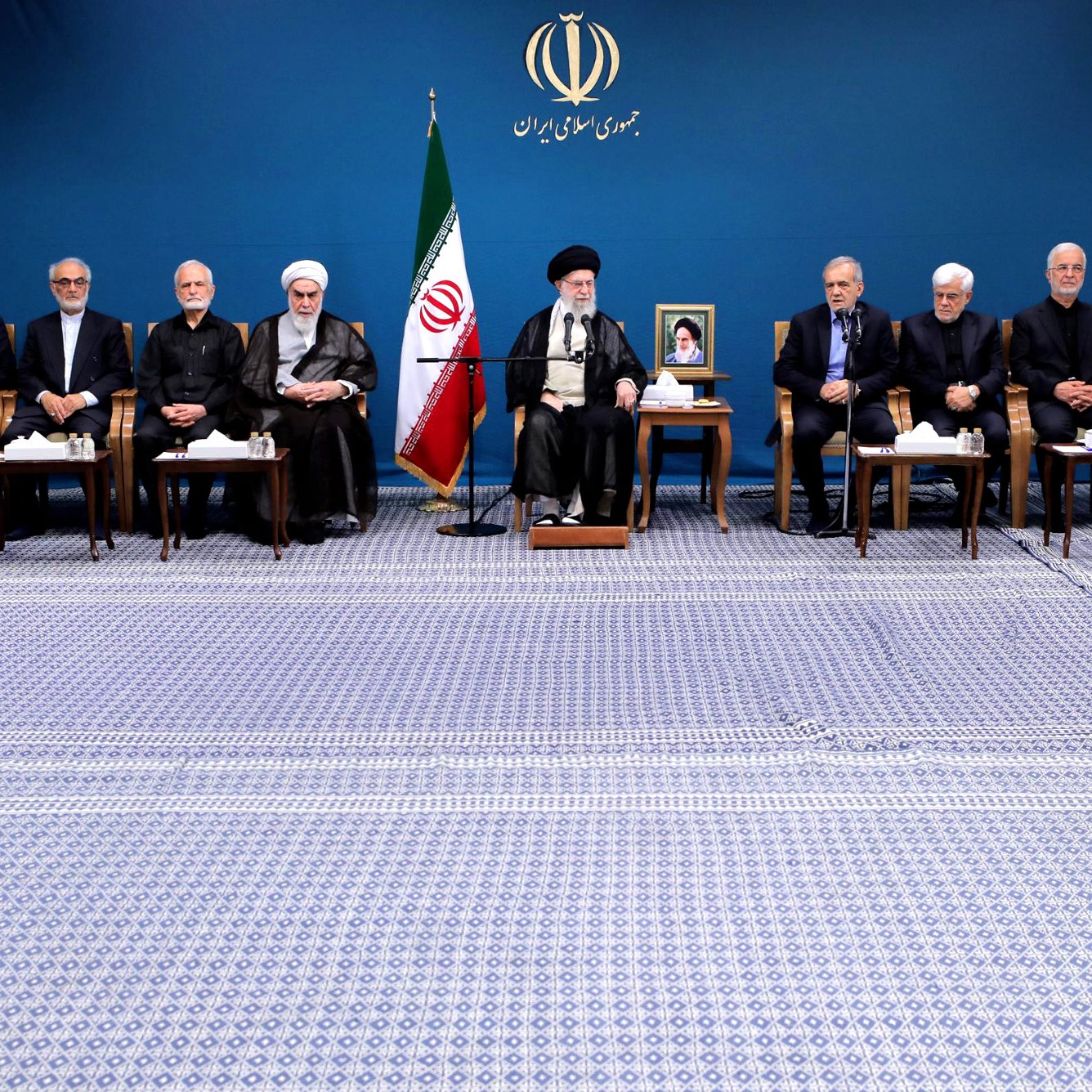 L'image montre un groupe de personnes assises en rangée sur un tapis, dans un environnement formel. En arrière-plan, il y a un drapeau iranien et un portrait d'une figure importante, probablement un leader. Les participants semblent être des hommes en costume sombre et en tenue religieuse, certains portant des turbans. Ils semblent engagés dans une discussion ou une réunion officielle. L'ambiance est solennelle et professionnelle.