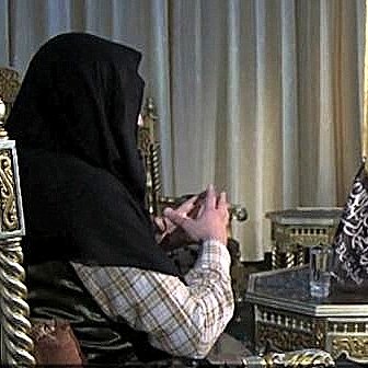 الصورة تظهر مشهداً بين شخصين يجلسان على كراسي مزخرفة. الشخص في الجهة اليسرى يرتدي ملابس تقليدية ويحمل بيديه بعض الأشياء. بينما الشخص في الجهة اليمنى يبدو أنه يقرا من كتاب أو ملاحظات. الخلفية تحتوي على زينة تقليدية، مع إضاءة ناعمة تعكس أجواء الحوار.