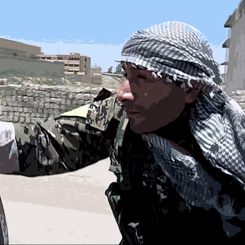 La imagen muestra a un hombre vestido con un uniforme militar y una kefía (una especie de pañuelo) en la cabeza. Él parece estar en un entorno urbano, posiblemente en un área de conflicto. El fondo incluye edificios y una zona que parece abandonada o dañada. El hombre tiene una expresión concentrada y parece estar en alerta, posiblemente observando o preparándose para algo en el entorno que lo rodea.