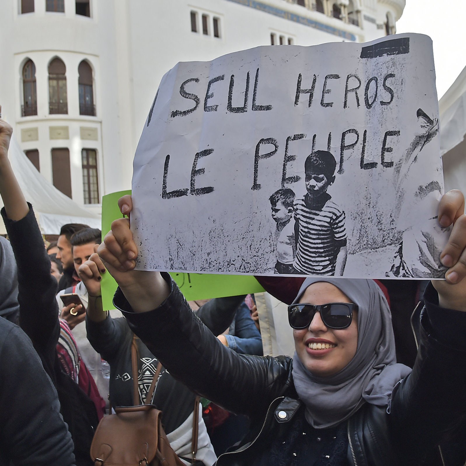 در این تصویر، تعدادی از مردم در یک تجمع حضور دارند. فردی جلوی دوربین ایستاده و یک پلاکارد بزرگ را در دست دارد که روی آن نوشته «SEUL HÉROS LE PEUPLE؟» به معنای «تنها قهرمان مردم؟» دیده می‌شود. فردی که پلاکارد را در دست دارد، عینک دودی به چشم دارد و روسری به سر دارد. اطراف او جمعیت متنوعی از افراد قابل مشاهده است که احتمالاً در حال مشارکت در یک رویداد اجتماعی یا اعتراض هستند. پس‌زمینه تصویر شامل سازه‌های معماری و چادرهایی است که به نظر می‌رسد برای این تجمع برپا شده‌اند.