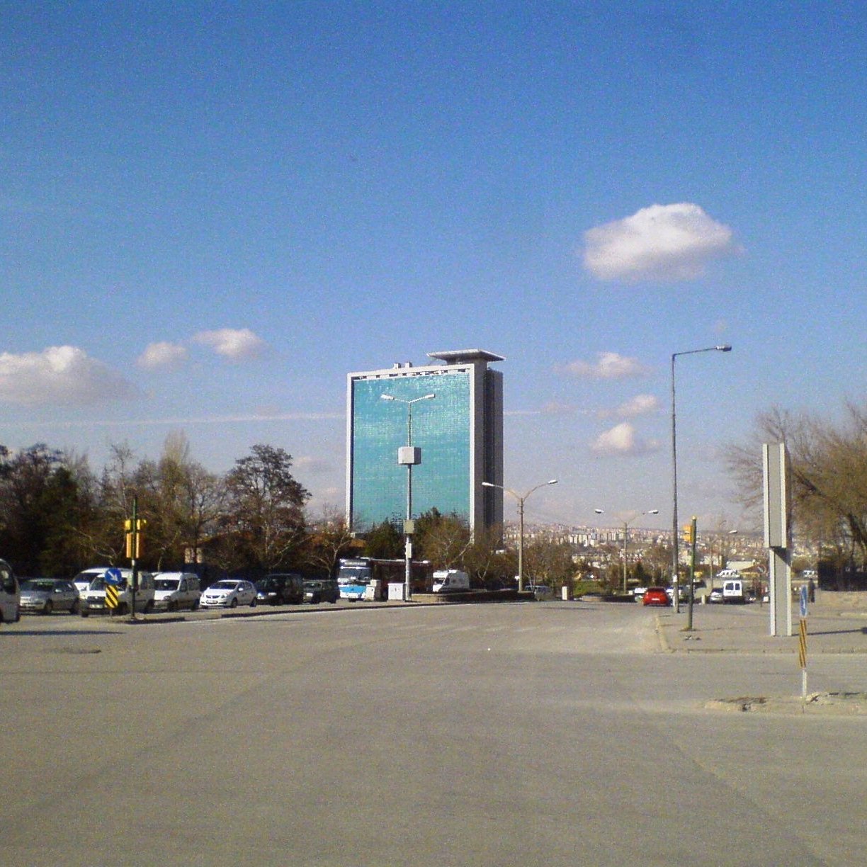 L'image montre une vue d'une rue avec un bâtiment moderne en arrière-plan. Ce bâtiment, de grande hauteur, a une façade en verre qui reflète le ciel. Le ciel est clair avec quelques nuages dispersés. On peut voir des voitures garées et circulant sur la route, ainsi que des arbres sur le côté. L'ensemble donne une impression de ville moderne et animée.