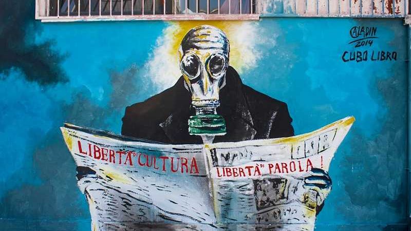 L'immagine rappresenta un murale che ritrae una figura umana con una maschera antigas, seduta e intenta a leggere un quotidiano. Il soggetto indossa un abbigliamento scuro e la scena è caratterizzata da una palette di colori vivaci, con sfumature di blu e bianco sullo sfondo. Le parole "LIBERTÀ", "CULTURA" e "LIBERTÀ di PAROLA!" sono scritte con un carattere forte e chiaro, sottolineando un messaggio di libertà e critica sociale. La data "2014" indica quando è stata realizzata l'opera, con il riferimento a "Cuba Libera" che suggerisce un legame con temi di libertà e diritti civili.