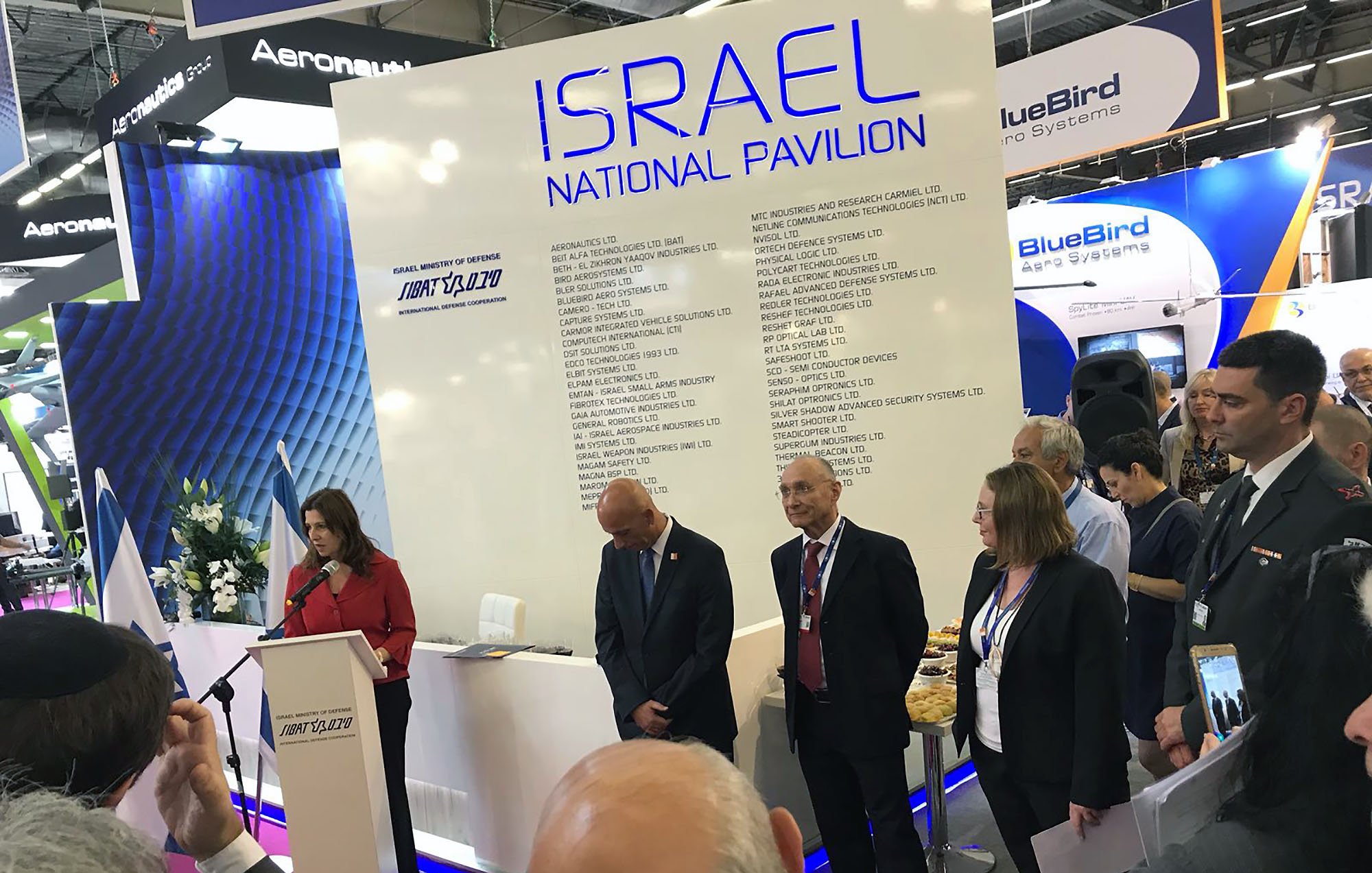The image shows a gathering at the Israel National Pavilion at an event or exhibition. A woman is speaking at a podium, while several individuals stand beside her, including a man in a suit and others who appear to be attentive to her speech. The background features a large display with the words “ISRAEL NATIONAL PAVILION” and various company names listed. The atmosphere seems formal, with an audience present, possibly indicating a business or governmental event.