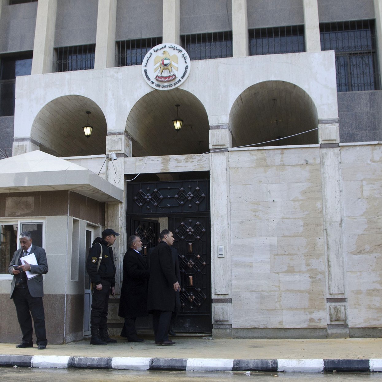 تصویر نشان‌دهندهٔ یک ساختمان دولتی است که به نظر می‌رسد در یک کشور عربی قرار دارد. در جلوی ساختمان، شبکه‌ای از ستون‌ها و پنجره‌ها وجود دارد و بر روی دیوار ورودی، نشان یا علامت مشخصی نصب شده است. چند نفر در جلوی ساختمان ایستاده‌اند؛ یکی در حال نصب علامت بر روی دیوار و دو نفر دیگر در حال گفتگو به نظر می‌رسند. همچنین، یک نگهبان در کنار ورودی قابل مشاهده است. زمینهٔ تصویر نیز آسفالت و خط‌کشی خیابان است.