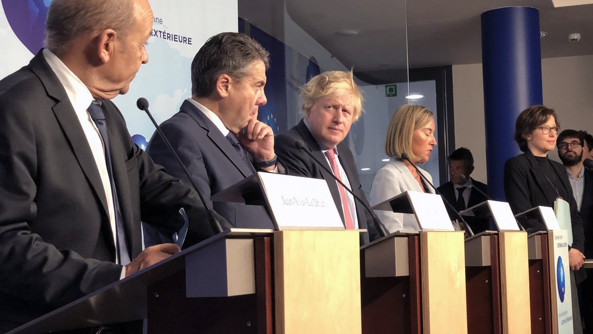 L'image montre une conférence de presse avec plusieurs personnes debout derrière des pupitres. Parmi eux, on peut voir des hommes en costume et une femme en tenue formelle. Ils semblent être en train de discuter ou d'écouter des questions, avec une atmosphère sérieuse. L'arrière-plan présente des éléments visuels représentant des thèmes internationaux, comme des logos ou des symboles géographiques.