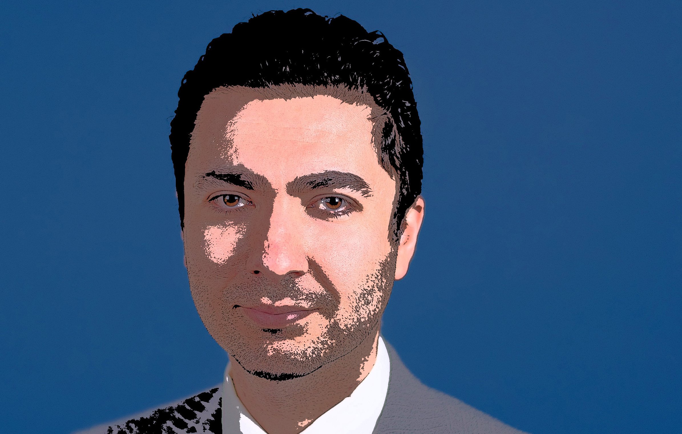 الصورة تظهر شخصاً يرتدي بدلة رسمية باللون الرمادي، مع قميص أبيض وربطة عنق زرقاء. الخلفية ذات لون أزرق، ويبدو على وجهه تعبير واثق. الشعر داكن وممشط بشكل مرتب.