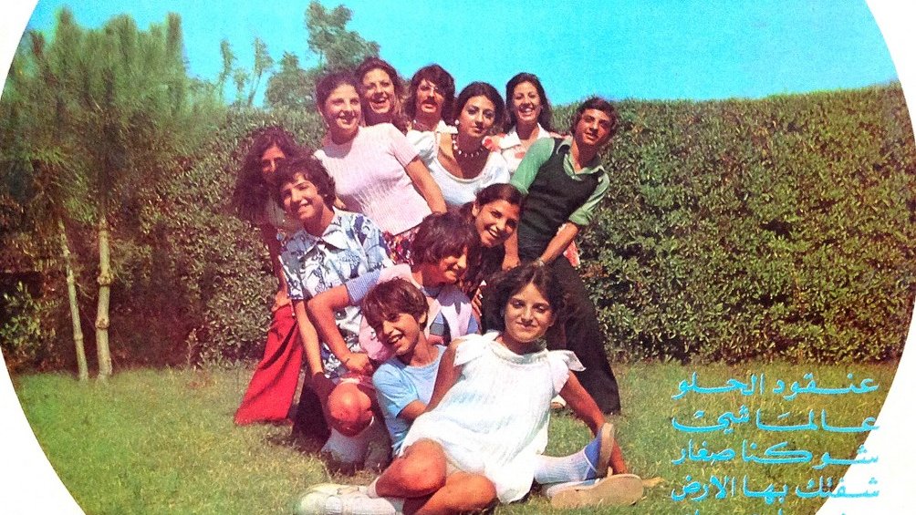 L'image représente une couverture de disque avec un groupe de personnes posant ensemble à l'extérieur, probablement dans un jardin. Ils semblent s'amuser et sourire, créant une ambiance joyeuse. Le nom du groupe, "BANDALY", et le titre "The Merry Family" sont présentés en bas, avec des inscriptions en arabe. En arrière-plan, on peut voir des arbres et une haie. L'ensemble évoque une atmosphère festive et conviviale.