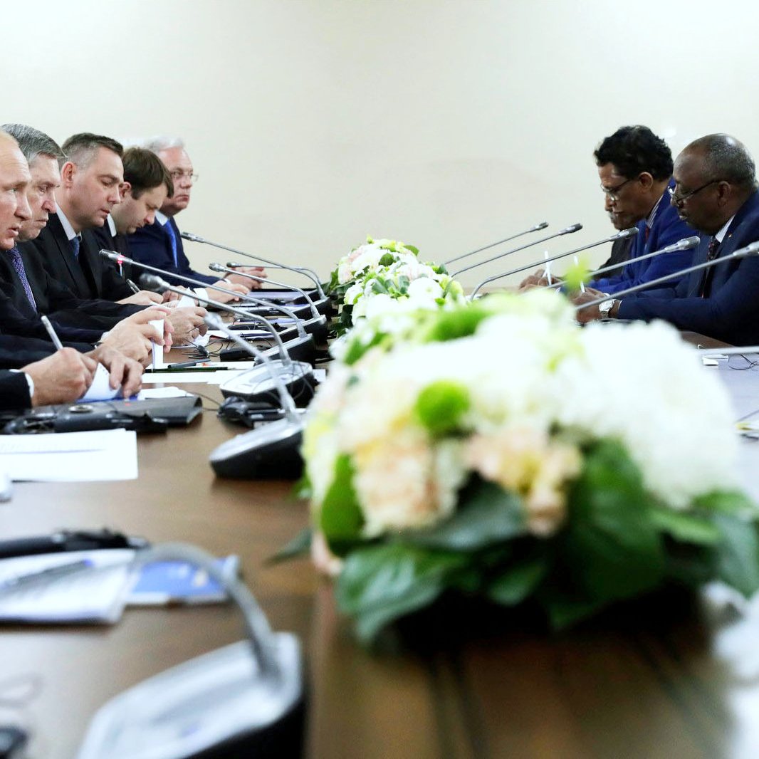 La imagen muestra una reunión formal en una sala con una mesa larga. Hay varias personas sentadas a ambos lados de la mesa, concentradas en la conversación. En el centro de la mesa, hay un arreglo floral decorativo. Las personas parecen estar intercambiando ideas, y algunos están tomando notas. El ambiente es serio y profesional.