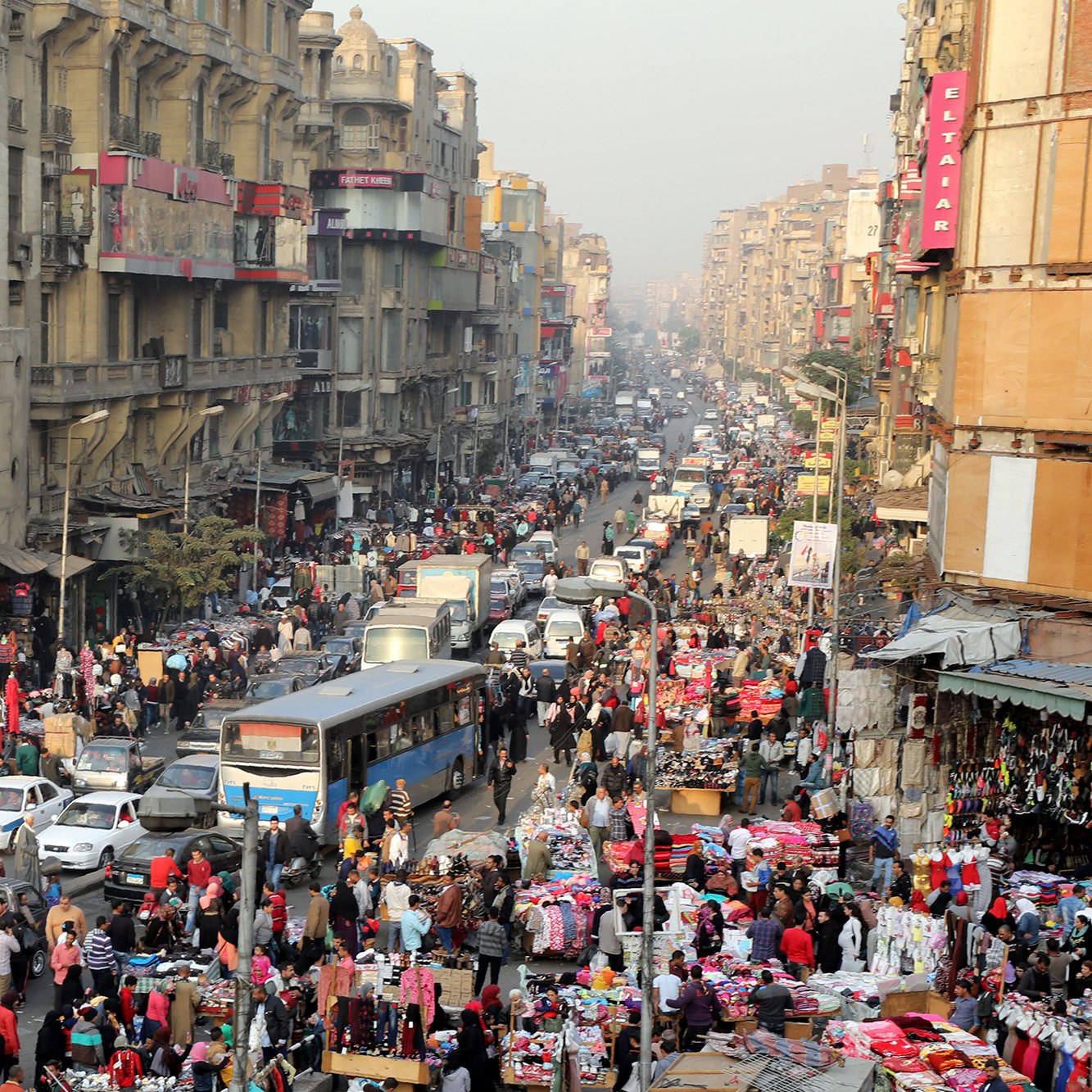 L'image montre une scène animée d'un marché urbain bondé, probablement dans une grande ville. On peut voir des foules de personnes se mêlant aux voitures dans une rue étroite. Les étals colorés sont remplis de divers produits, tels que des vêtements et des accessoires. Les bâtiments environnants semblent anciens et plusieurs niveaux sont visibles, avec des enseignes visibles. L'atmosphère paraît vivante et dynamique, typique des marchés populaires dans les grandes agglomérations.
