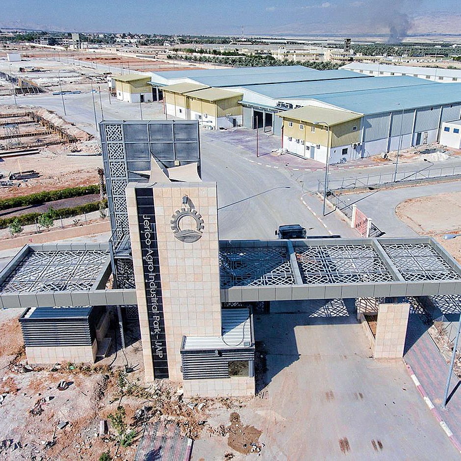 The image shows a large industrial complex with a modern entrance gate. The entrance features a prominent archway or facade, likely decorated with geometric patterns or designs. Surrounding the gate, there are several buildings, and the area appears to be part of a developing industrial site, as indicated by construction materials and equipment visible in the background. The landscape is predominantly dry and arid, with sparse greenery, and you can see a distant horizon with some smoke rising, suggesting nearby activity or construction. Solar panels are also visible on the ground, indicating a focus on sustainable energy.