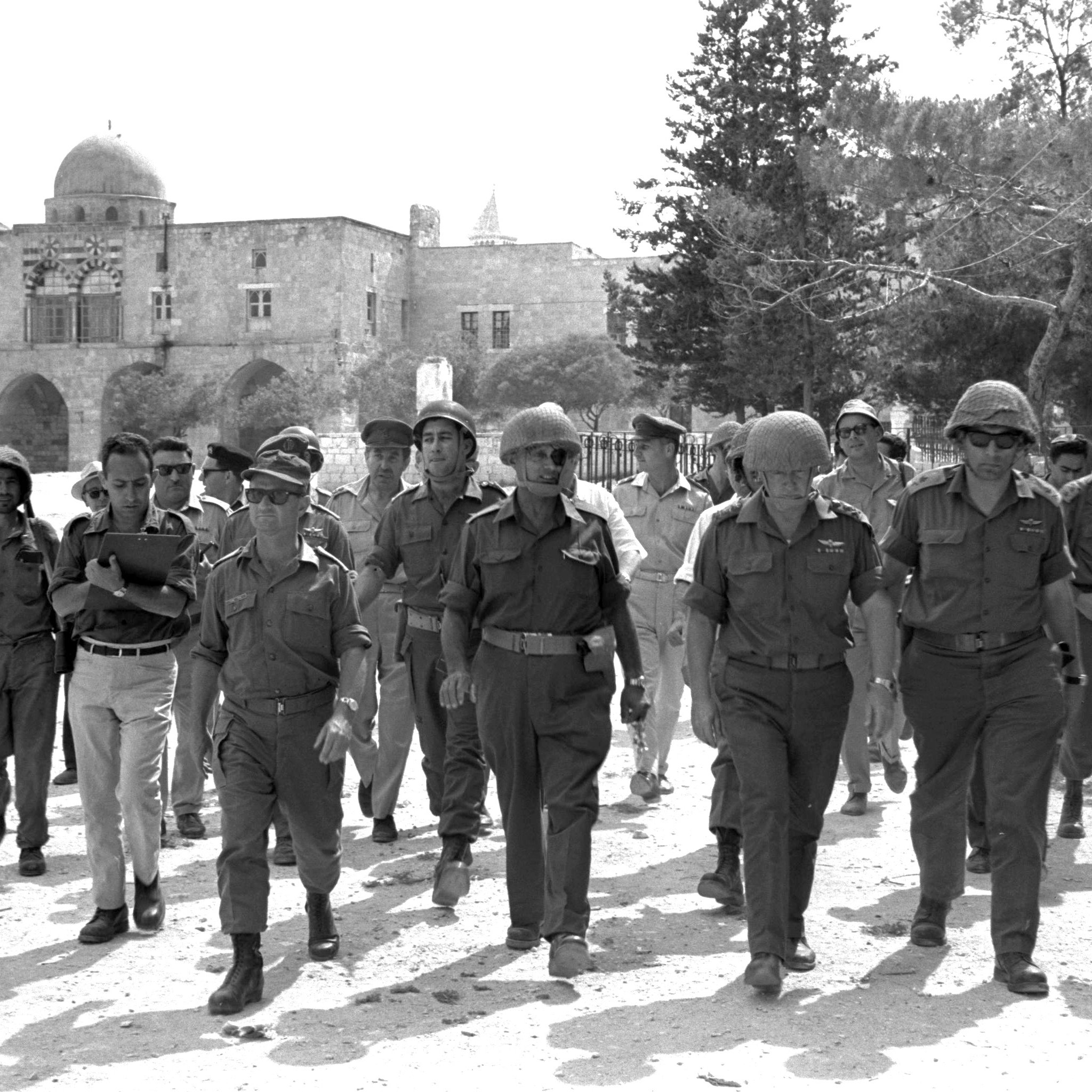 در این تصویر، گروهی از نظامیان و مأموران امنیتی در حال راه رفتن به سمت دوربین هستند. آن‌ها لباس‌های نظامی و کلاه‌های مخصوص به سر دارند. پس‌زمینه تصویر، شامل ساختمان‌های سنگی قدیمی و برخی درختان است. به نظر می‌رسد این تصویر در یک منطقه تاریخی یا باستانی گرفته شده باشد. نور خورشید به وضوح بر روی صحنه می‌تابد و فضایی گرم به تصویر می‌بخشد.