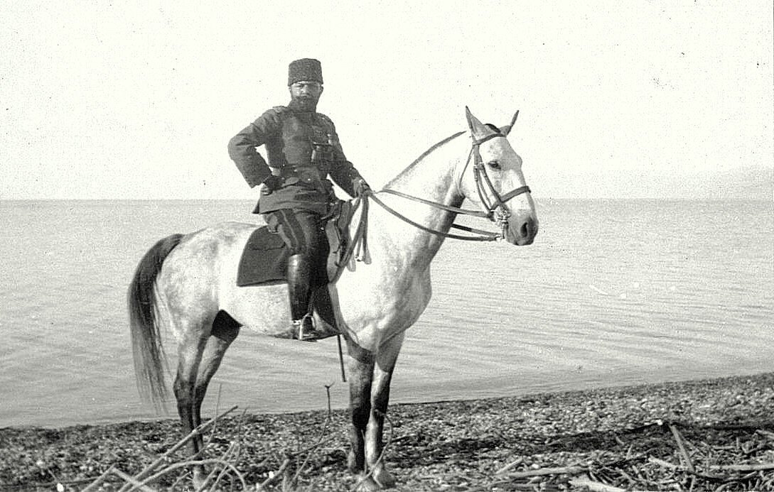 در تصویر، یک مرد uniform پوشیده بر روی یک اسب سفید ایستاده است. او در کنار آب قرار دارد و زمینه‌ای آرام و طبیعی دیده می‌شود. لباس او با جزئیات مشخصی همراه است و حالتی رسمی دارد. اسب نیز با ظاهر زیبا و شکیل خود توجه را جلب می‌کند. این صحنه به نظر می‌رسد در زمان گذشته و در مکانی نزدیک به دریا یا دریاچه ثبت شده باشد.