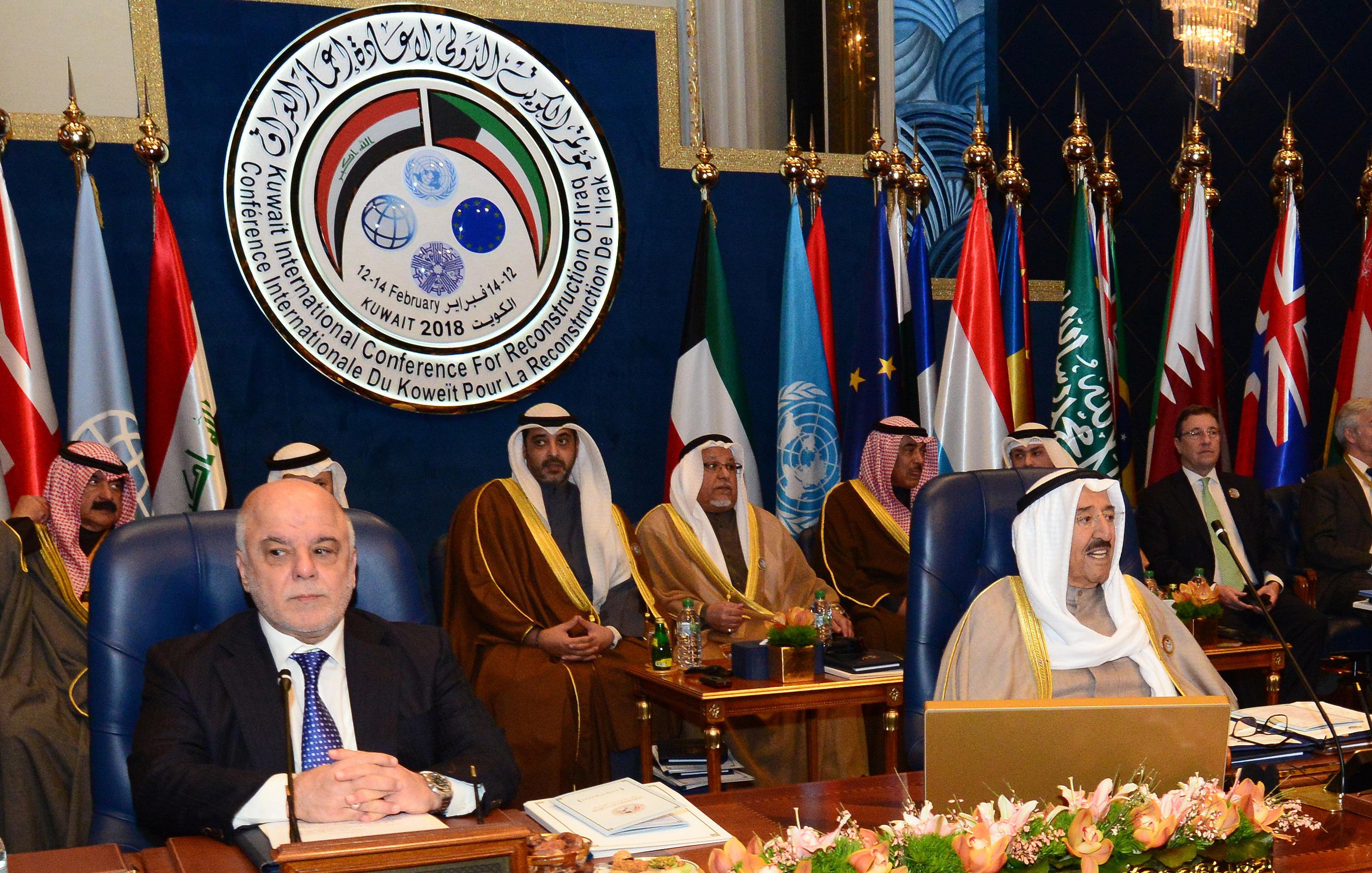 The image depicts a formal conference setting, featuring several dignitaries sitting in a room with flags from various nations displayed prominently in the background. The attendees are dressed in traditional attire, indicating a Middle Eastern cultural context. A large emblem or logo is visible above them, likely representing the conference's purpose, which involves international cooperation or reconstruction efforts. Among the seated figures, one appears to be a prominent leader or official, positioned at the forefront of the image. The overall atmosphere suggests a significant meeting aimed at addressing important regional or global issues.