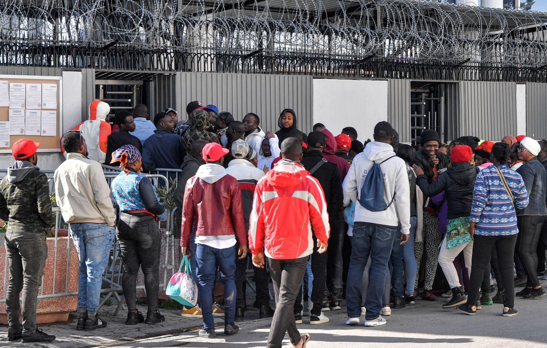 L'image montre un groupe de personnes rassemblées devant une clôture avec des barbelés. Elles semblent attendre ou essayer d'entrer dans un bâtiment. Certaines personnes portent des vêtements colorés, comme des vestes et des chapeaux rouges. Il y a une certaine densité dans la foule, suggérant une situation de rassemblement ou de file d'attente. Le mur en arrière-plan comporte des affichages et il y a une ambiance de tension ou d'importance autour de l'endroit où se trouvent les personnes.
