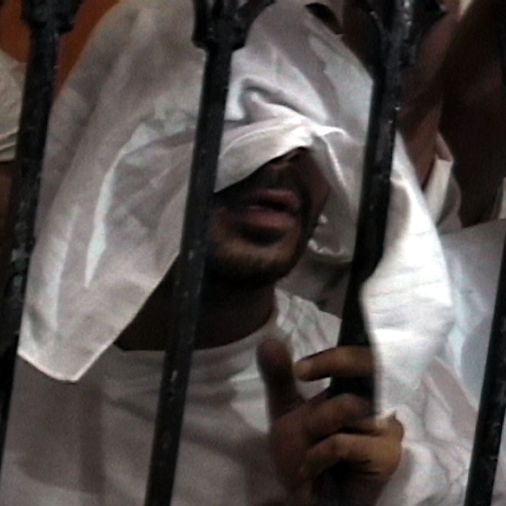L'image montre une personne avec un regard partiellement caché par un tissu blanc. Elle se trouve derrière des barreaux, ce qui suggère une situation de confinement ou de détention. Les mains de la personne sont visibles, tenant les barreaux, et le fond de l'image semble flou, mettant l'accent sur la figure centrale. L'atmosphère de l'image peut évoquer des sentiments d'isolement ou de détresse.