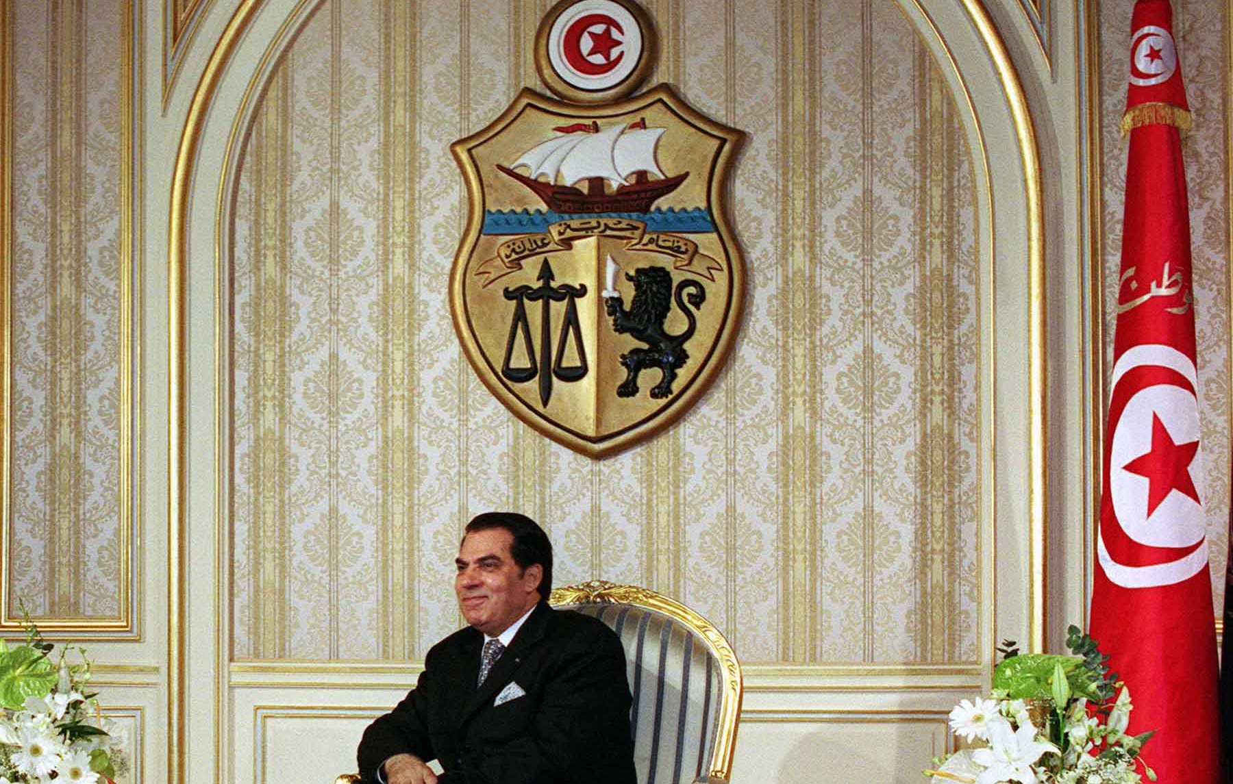 L'image montre un homme assis sur une chaise élégante dans un environnement formel. Il porte un costume sombre et semble très confortable. À l'arrière-plan, on peut voir un grand blason tunisien ainsi qu'un drapeau de la Tunisie, ce qui indique qu'il s'agit d'un cadre officiel. Des fleurs blanches sont placées sur des tables basses de chaque côté de lui, ajoutant une touche de sophistication à la scène. Les murs sont décorés de manière riche, ce qui renforce le caractère solennel de l'environnement.