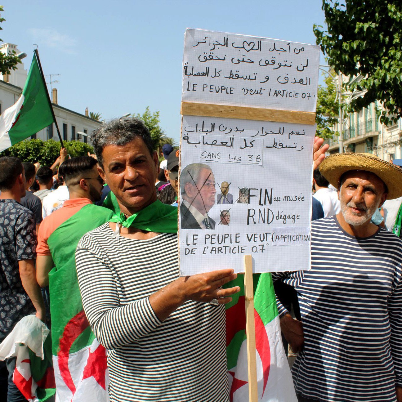 تظهر في الصورة مجموعة من الأشخاص في تجمع أو مظاهرة. يحمل بعضهم لافتات تتضمن شعارات سياسية تتعلق بالمطالب الشعبية في الجزائر. يرتدي المشاركون ملابس بألوان متنوعة، ويبدو أن هناك استخداماً للعلم الجزائري. المشهد يعكس روح الحماس والتعبير عن المطالب الجماهيرية.