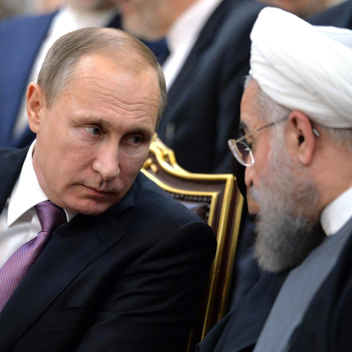 The image depicts two men engaged in a conversation. One is seated with a serious expression, wearing a dark suit and a light-colored tie. The other man, also seated, has a beard and wears traditional clerical attire, including a white turban. They appear to be in a formal setting, possibly during a meeting or event, with other individuals visible in the background. The focus is on their interaction and expressions.