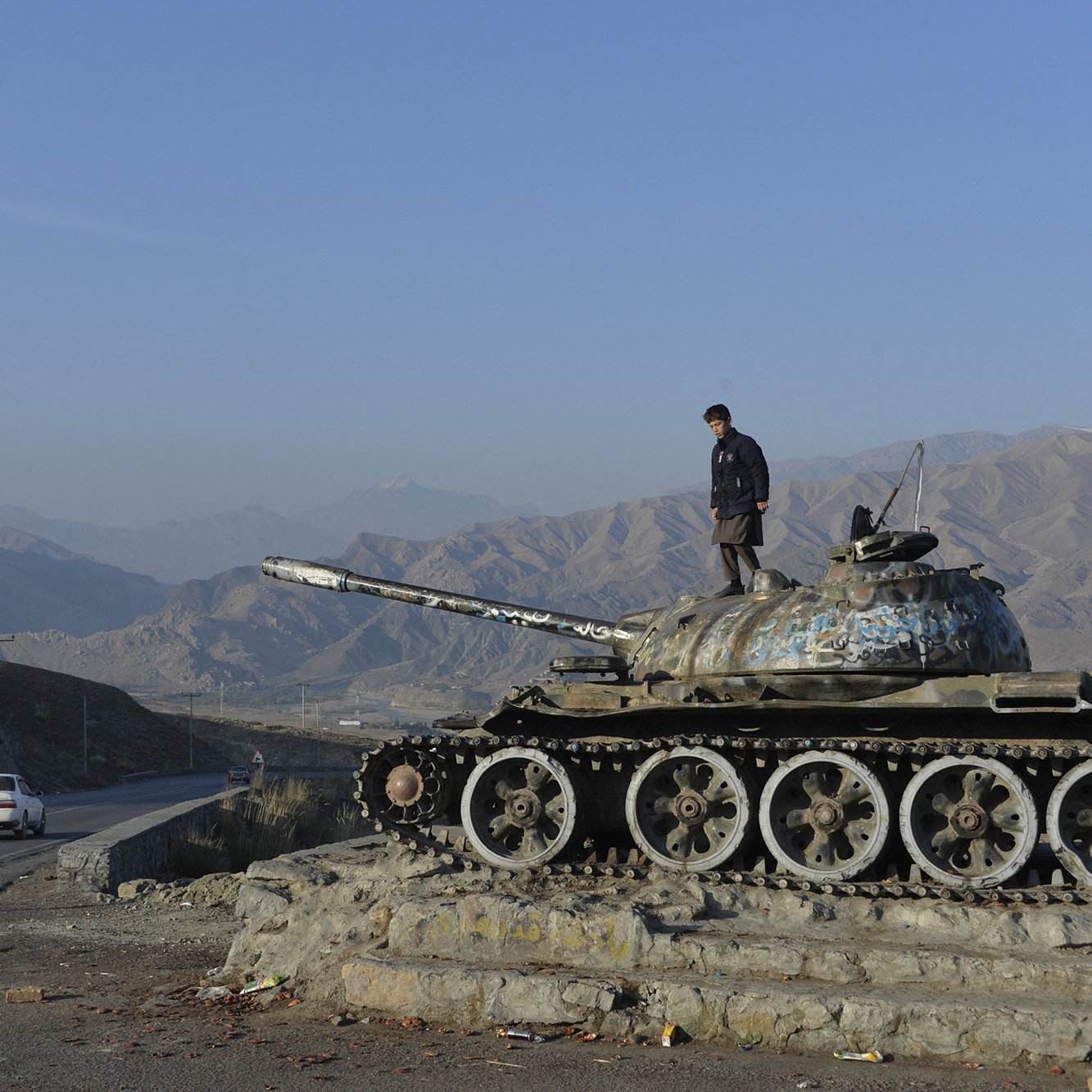 La imagen muestra a un joven de pie sobre un tanque de guerra, que parece estar fuera de uso. El tanque tiene un acabado desgastado y está cubierto de óxido y pintura descascarada. Al fondo, se pueden ver montañas cubiertas de vegetación y un cielo despejado. A un lado, hay una carretera donde se observa un automóvil blanco en movimiento. La escena transmite una sensación de desolación y el contraste entre la guerra y la paz.
