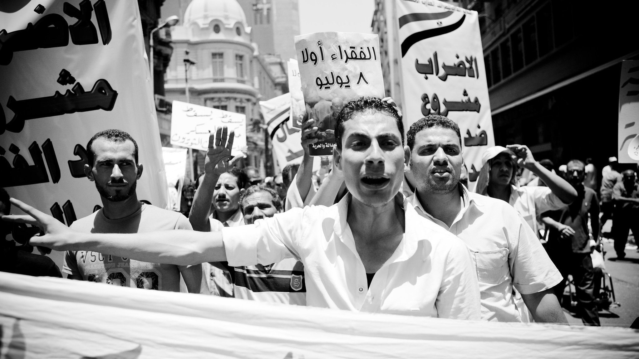 The image depicts a black and white scene of a protest or demonstration. A group of people is marching together, holding banners and signs. One person in the foreground appears to be passionately speaking or chanting, while others raise their fists or shout in the background. The signs seem to convey messages related to social or political issues, with at least one sign written in Arabic. The overall atmosphere suggests urgency and activism, capturing a moment of collective expression and dissent.