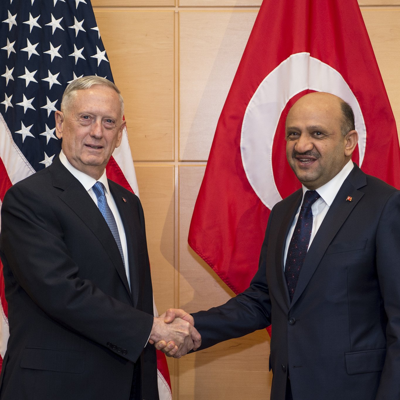 در این تصویر، دو مرد در حال دست دادن هستند. در پس‌زمینه، پرچم‌های ایالات متحده و ترکیه دیده می‌شوند. یکی از مردان، با موهای خاکستری و کت و شلوار تیره، به نظر می‌رسد یک مقام آمریکایی باشد. مرد دیگر نیز با کت و شلوار تیره و کراواتی با طرحی خاص، نماینده ترکیه به نظر می‌رسد. این تصویر به نوعی نشان‌دهنده همکاری یا مذاکرات بین دو کشور است.