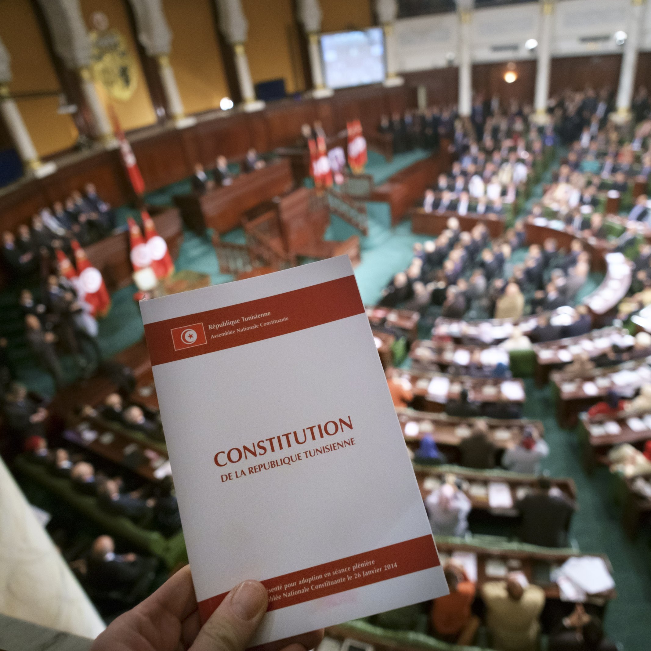 The image shows a parliamentary session in Tunisia, with a focus on a hand holding a book titled "Constitution de la République Tunisienne" (Constitution of the Tunisian Republic). In the background, a large assembly of representatives is seated in a formal, legislative setting, likely discussing or voting on matters related to the constitution or other important legislative issues. The architecture of the chamber features ornate details, and national flags are visible, emphasizing the official nature of the event.