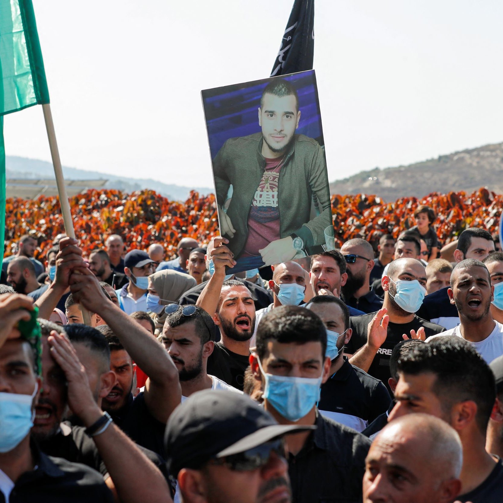 الصورة تُظهر مجموعة من المتظاهرين في تجمع حاشد، يحمل بعضهم لافتات وأعلام. وسط الحشد، يوجد شخص يحمل صورة لرجلٍ يبدو أنه شخصية مهمة أو رمزية بالنسبة لهم. يرتدي الكثير من المتظاهرين الكمامات، ويبدو أن المظاهرة تتم في منطقة مفتوحة، مع خلفية تضم تلالًا وأشجار. تُعبر الأجواء عن الحماس والتجمع الجماهيري.