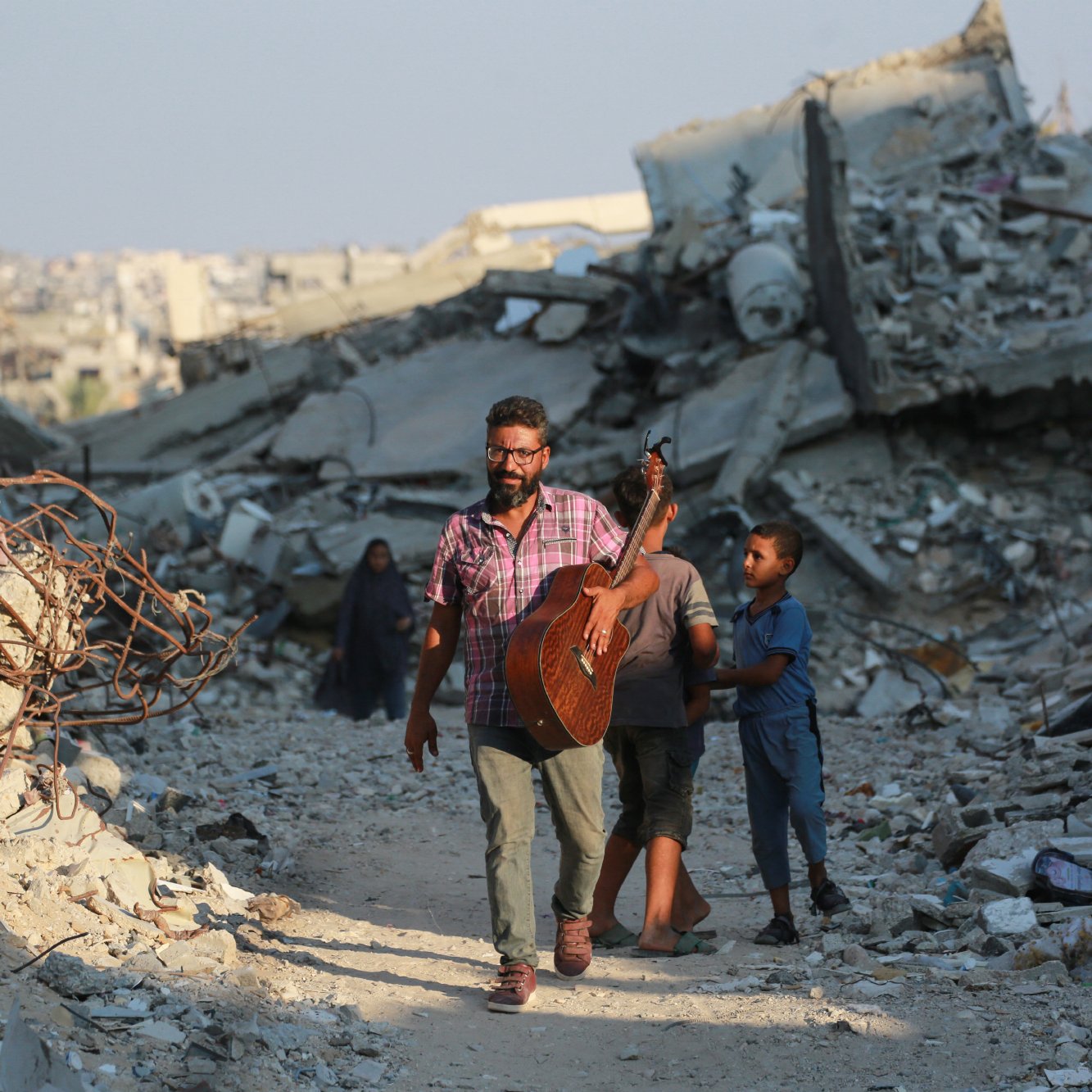 L'immagine mostra un uomo che cammina tra le macerie di un'area distrutta, portando una chitarra. Sembra essere felice mentre si muove, accompagnato da un bambino. Sullo sfondo ci sono molti detriti e edifici in rovina, che suggeriscono una situazione di distruzione. Un'altra figura, probabilmente una donna, è visibile più lontano tra le macerie. L'atmosfera trasmette un contrasto tra la musica e la devastazione circostante.