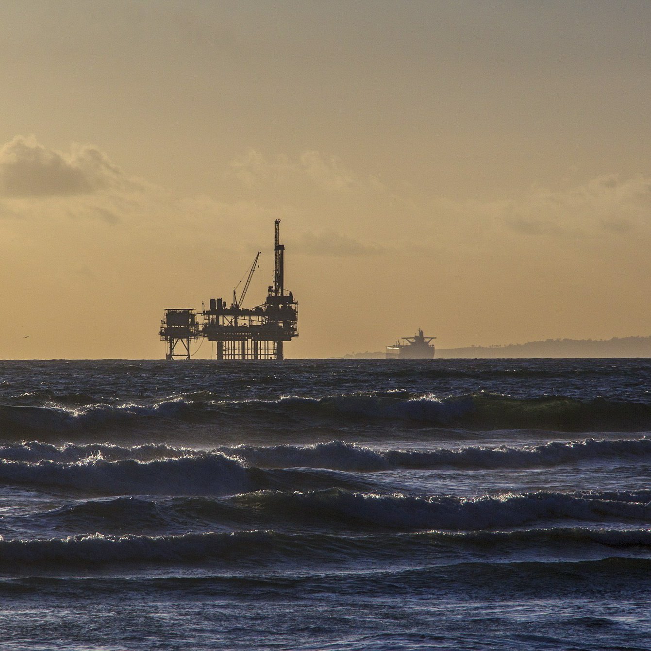The image shows a distant oil rig standing tall in the ocean during a sunset. The sky is illuminated with soft golden hues, creating a serene atmosphere. Waves roll gently in the foreground, and in the background, a cargo ship can be seen faintly on the horizon. The overall mood is calm and peaceful, with a blend of industrial and natural elements.