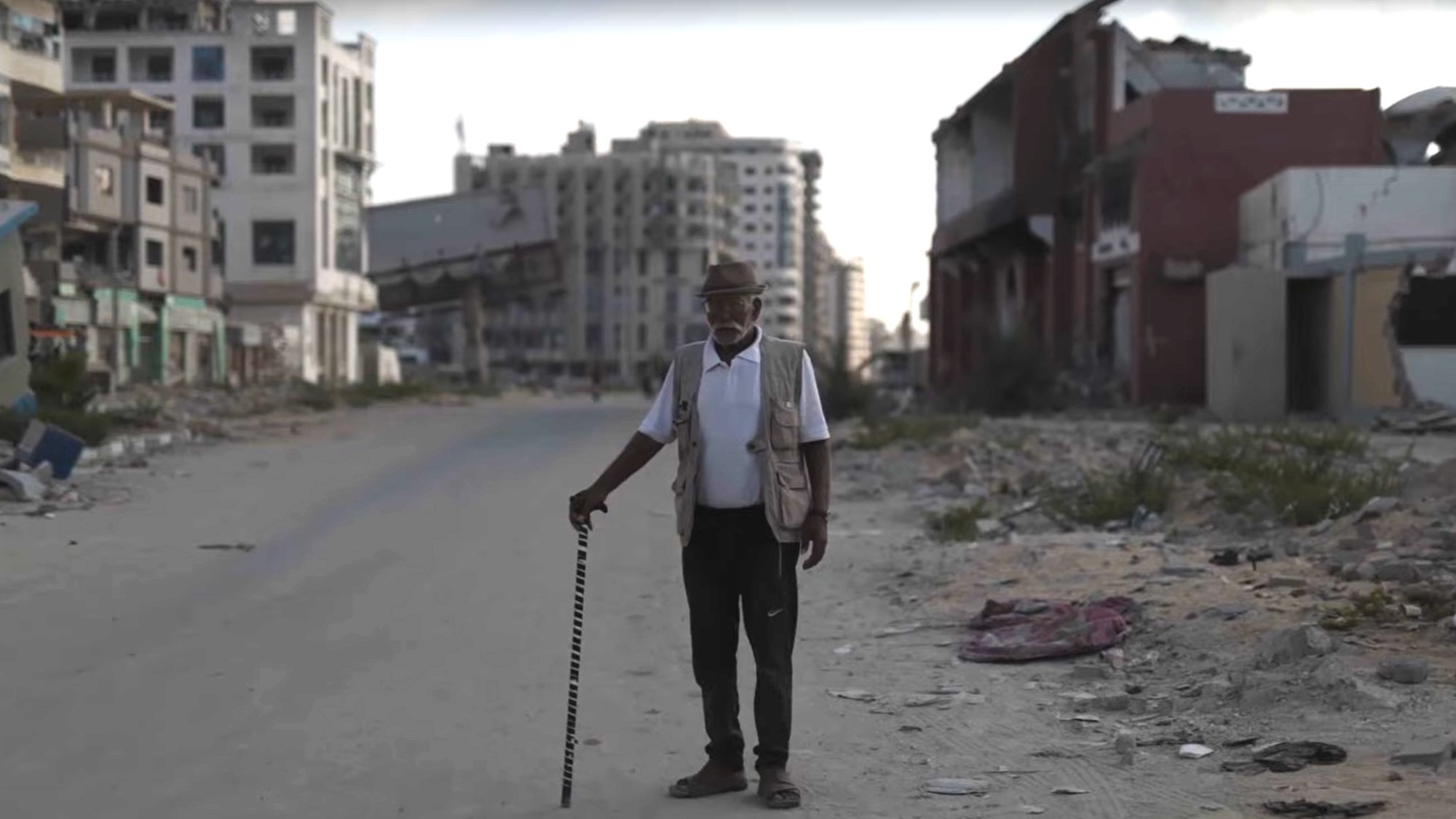 Un homme âgé avec une canne se tient sur une rue déserte, entouré de bâtiments en ruine.