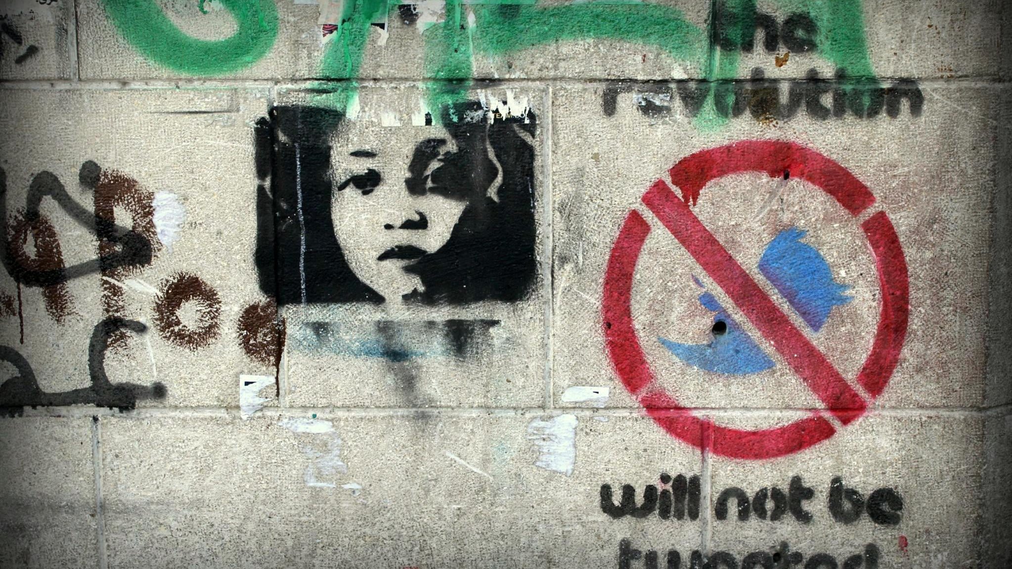 L'image présente un mur couvert de graffitis. On y voit un portrait en noir et blanc d'un enfant qui regarde vers l'observateur. À côté de ce portrait, il y a des inscriptions en couleur, dont l'une mentionne "the revolution" (la révolution) et une autre un symbole barré de l'oiseau bleu, accompagnée du texte "will not be tweeted" (ne sera pas tweeté). L'ensemble évoque des thèmes de révolte et de critique des réseaux sociaux.