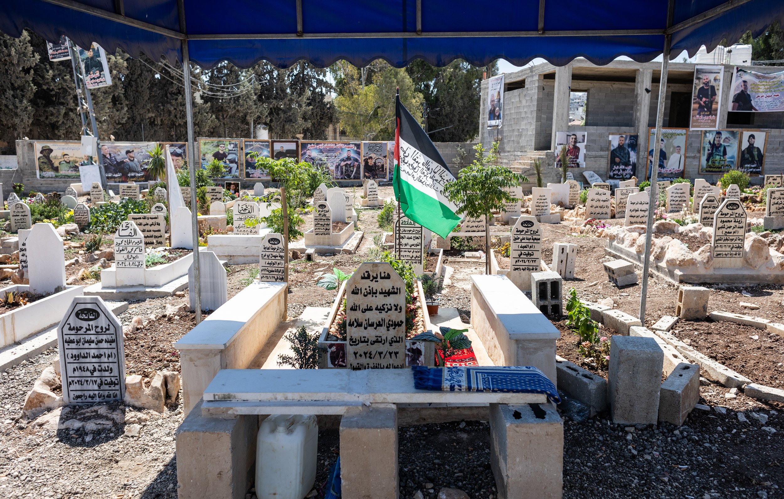 L'immagine mostra un cimitero all'aperto, con diverse tombe disposte in un'area ben curata. Le lapidi sono in varie posizioni e materiali, molte sono contrassegnate con iscrizioni in arabo. Ci sono anche alcune piante verdi e fiori, che aggiungono un tocco di vita al luogo. Al centro, si può vedere una bandiera con i colori della Palestina che sventola, e sullo sfondo ci sono delle fotografie appese, probabilmente in memoria di persone care. Il cimitero è coperto da una tettoia, creando un'area protetta. L'atmosfera è solenne e rispettosa.