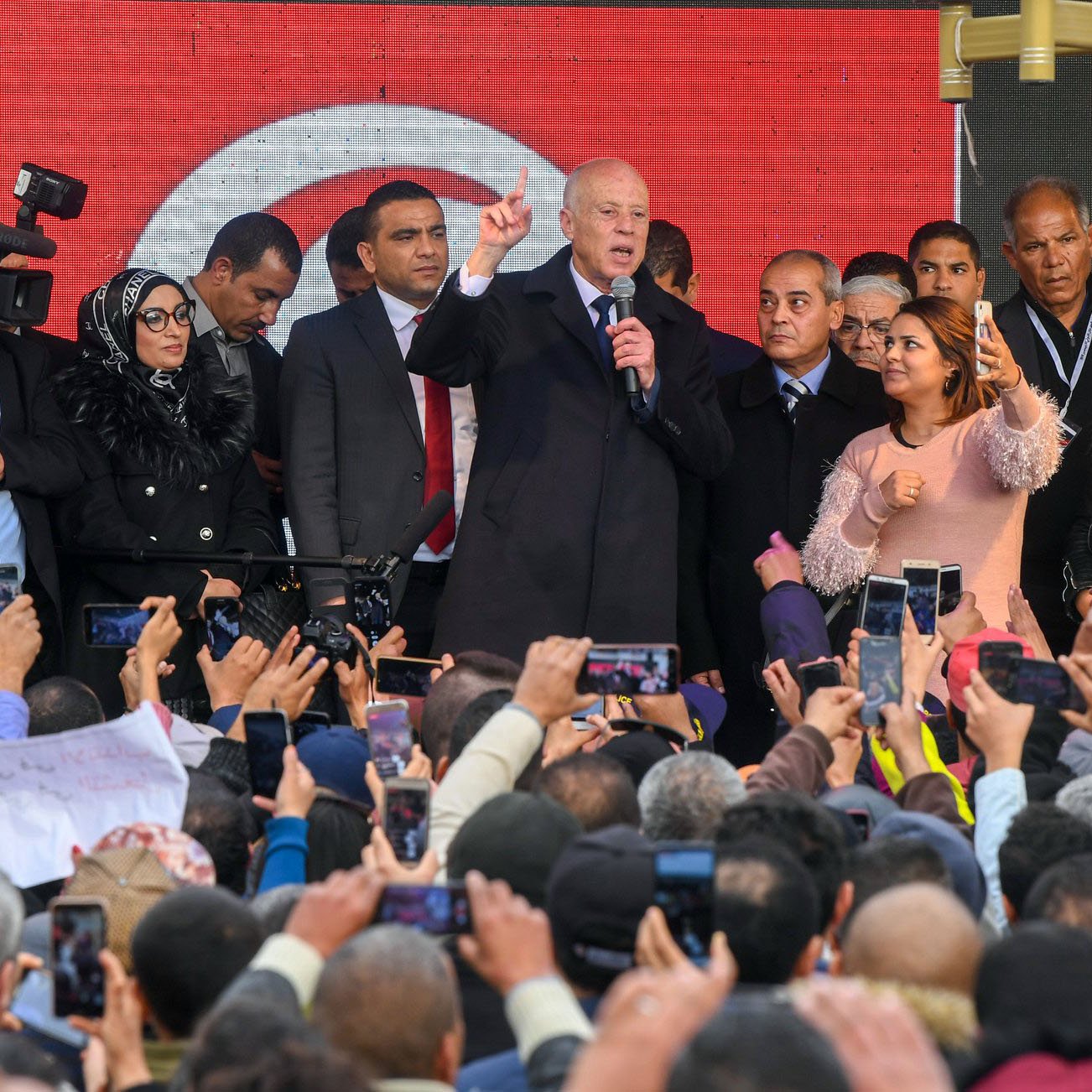 تظهر في الصورة حشود كبيرة من الناس تجمعوا في مكان عام. في المقدمة، يظهر شخص يقف على منصة ويتحدث، بينما يمسك العديد من الحضور هواتفهم المحمولة لتوثيق اللحظة. يحيط به مجموعة من الأشخاص، وبعضهم يبدو مشغولاً بالتقاط الصور أو الفيديو. خلف المنصة، توجد لافتة تحمل الألوان الوطنية. الأجواء توحي بالحماس والاهتمام من الحضور.