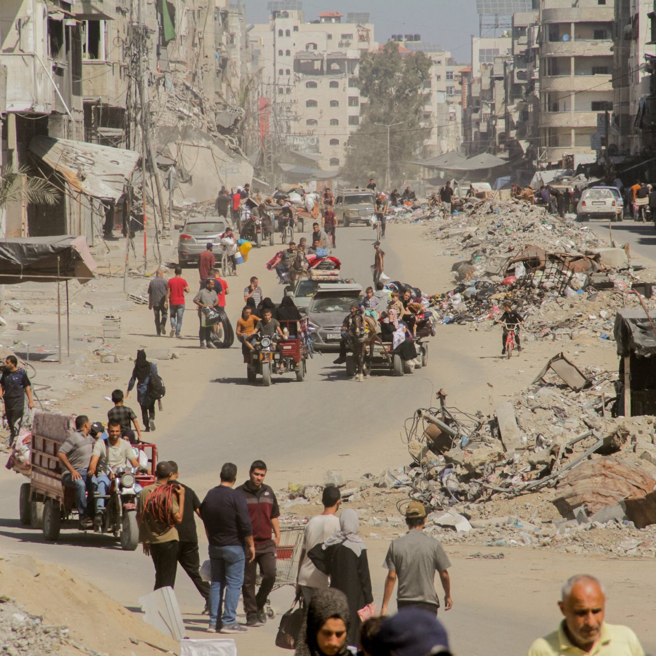 L'image montre une rue dévastée, probablement dans une zone de conflit. On peut voir des bâtiments en ruines de chaque côté de la voie, avec des débris éparpillés sur le sol. De nombreuses personnes, dont des familles et des individus, se déplacent sur la route. Certains utilisent des véhicules à trois roues, tandis que d'autres marchent à pied. L'atmosphère semble être celle d'une zone en reconstruction, avec beaucoup de poussière dans l'air et une ambiance de désolation.