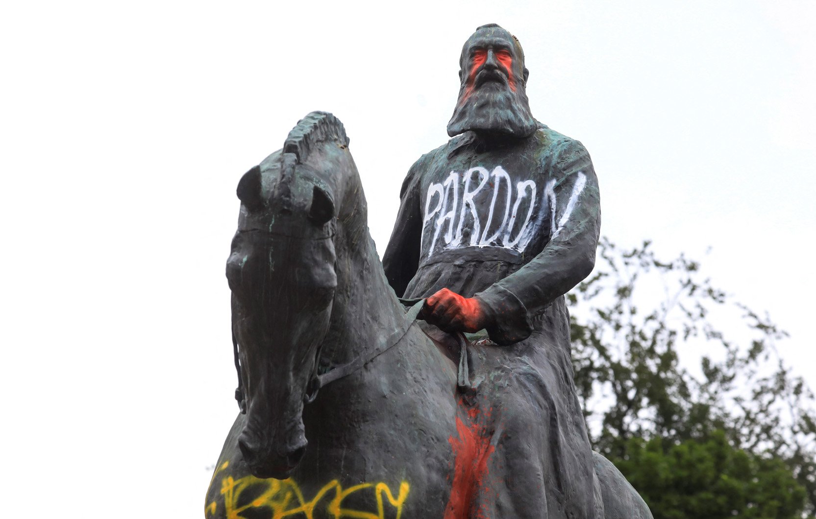 L'image montre une statue équestre d'un homme, probablement un personnage historique, qui porte des graffitis. Le mot "PARDON" est écrit en lettres blanches sur sa robe, tandis que d'autres mots, comme "RACIST", apparaissent en couleur sur le socle. La statue semble être recouverte de peinture, ce qui suggère un acte de protestation ou de critique. En arrière-plan, on peut voir un ciel nuageux et quelques arbres.