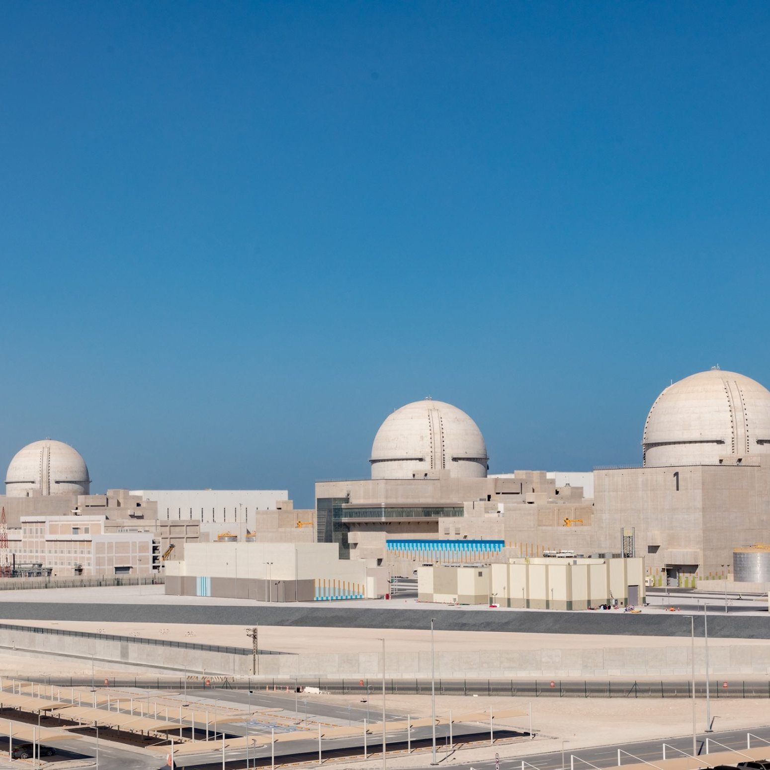 L'image montre une centrale nucléaire moderne avec plusieurs grands dômes blancs sur le toit. La structure est entourée d'un paysage clair, avec un ciel bleu sans nuages. À l'avant-plan, on peut voir des bâtiments de différentes tailles, ainsi qu'une zone de stationnement. L'ensemble de la scène dégage une impression de technologie avancée et de robustesse.