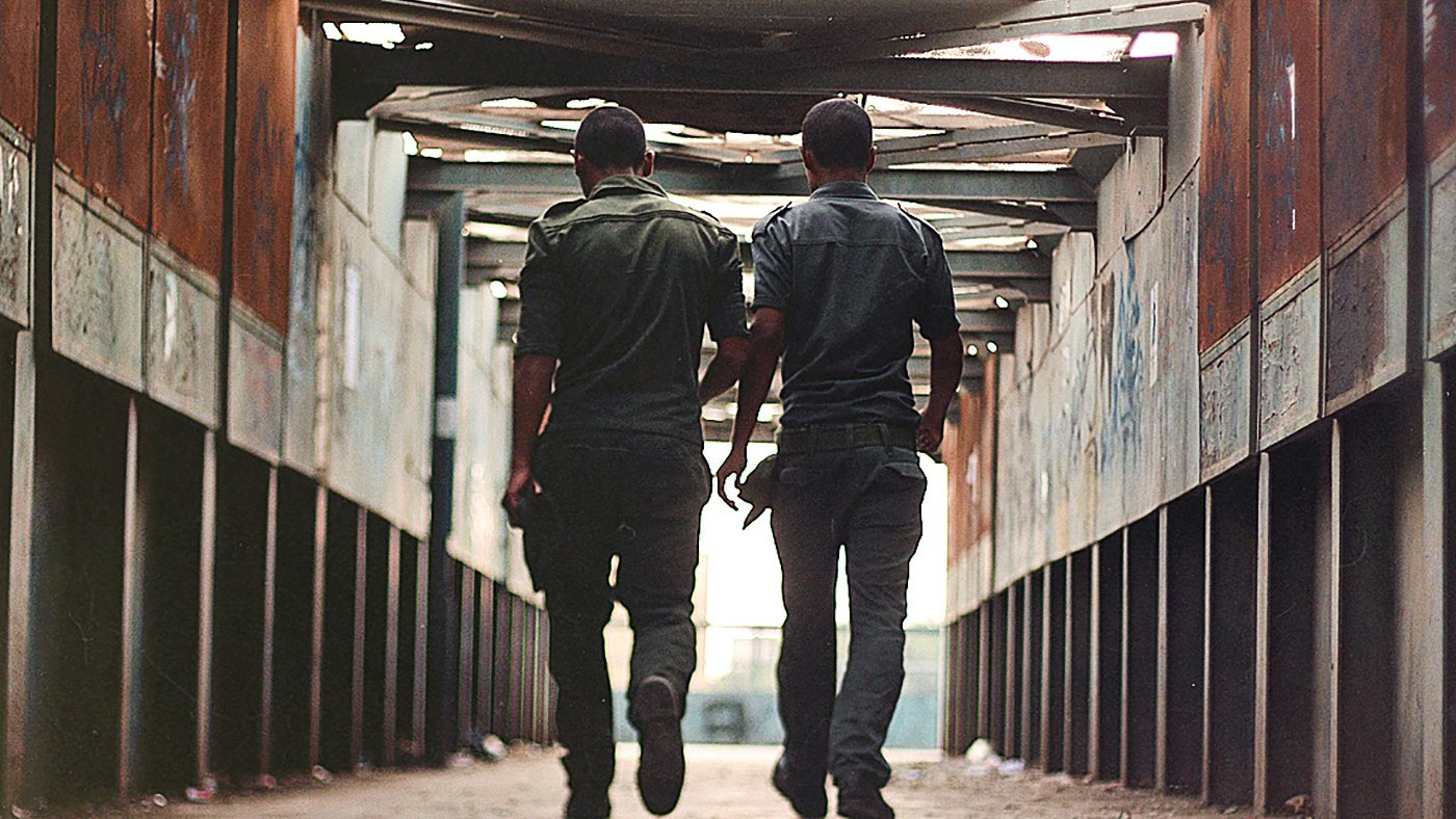 L'image montre deux hommes marchant côte à côte dans un passage couvert. Ils portent des uniformes de couleur sombre et se déplacent vers la sortie, avec leurs silhouettes se détachant contre la lumière. Les murs du passage semblent usés, avec des graffitis visibles, et le sol est pavé. L'atmosphère est à la fois urbaine et légèrement mélancolique, donnant une impression d'intimité entre les deux personnages.