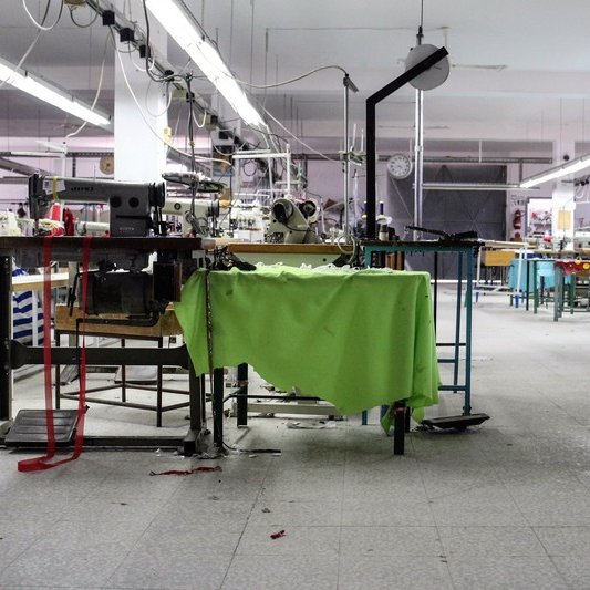 تظهر الصورة مشهدًا داخليًا لورشة خياطة. المساحة تبدو واسعة ومشرقة، حيث توجد آلات خياطة متعددة منتشرة في المكان. بعض الطاولات تحتوي على أقمشة ملونة، في حين أن العديد من المعدات والأدوات موجودة في حالة استخدام أو جاهزية. الأرضية مغطاة ببعض القصاصات من القماش، مما يشير إلى نشاط العمل. الإضاءة تأتي من الأنوار السقفية، وهناك ساعة على الحائط تدل على الوقت. يبدو أن الورشة تحتاج إلى بعض الترتيب.