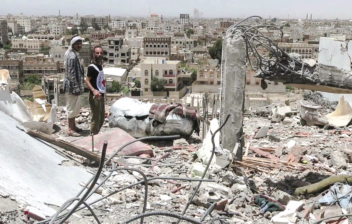 L'image montre deux hommes se tenant au milieu des décombres d'un bâtiment détruit, surplombant une ville en arrière-plan. Les structures environnantes semblent endommagées, avec de nombreux bâtiments partiellement effondrés et une apparence de désolation. Le ciel est clair, mais l'environnement porte des traces évidentes de conflit. Les hommes semblent examine la scène, témoignant d'une situation difficile et dévastatrice.