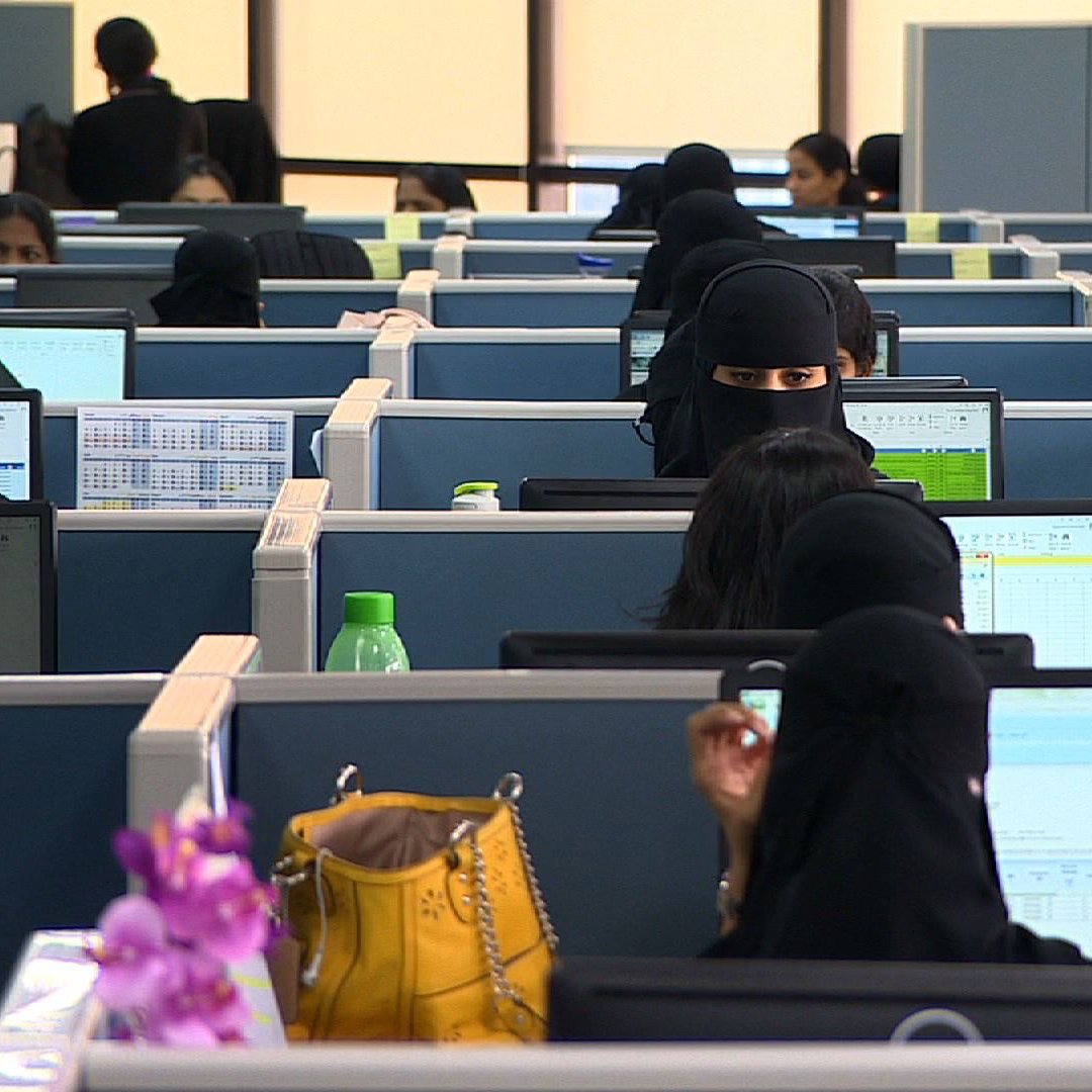 این تصویر یک محیط اداری را نشان می‌دهد که در آن تعدادی زن در حال کار هستند. بسیاری از این زنان از پوشش کامل استفاده می‌کنند و فقط چهره‌هایشان قابل مشاهده است. میزها به صورت ردیفی در کنار هم قرار دارند و هر نفر در حال کار با کامپیوتر است. در قسمت جلویی تصویر، چند شیء مانند بطری آب و یک کیف دیده می‌شود. فضای کلی تصویری از یک محیط کاری فعال و سازمان‌یافته است.