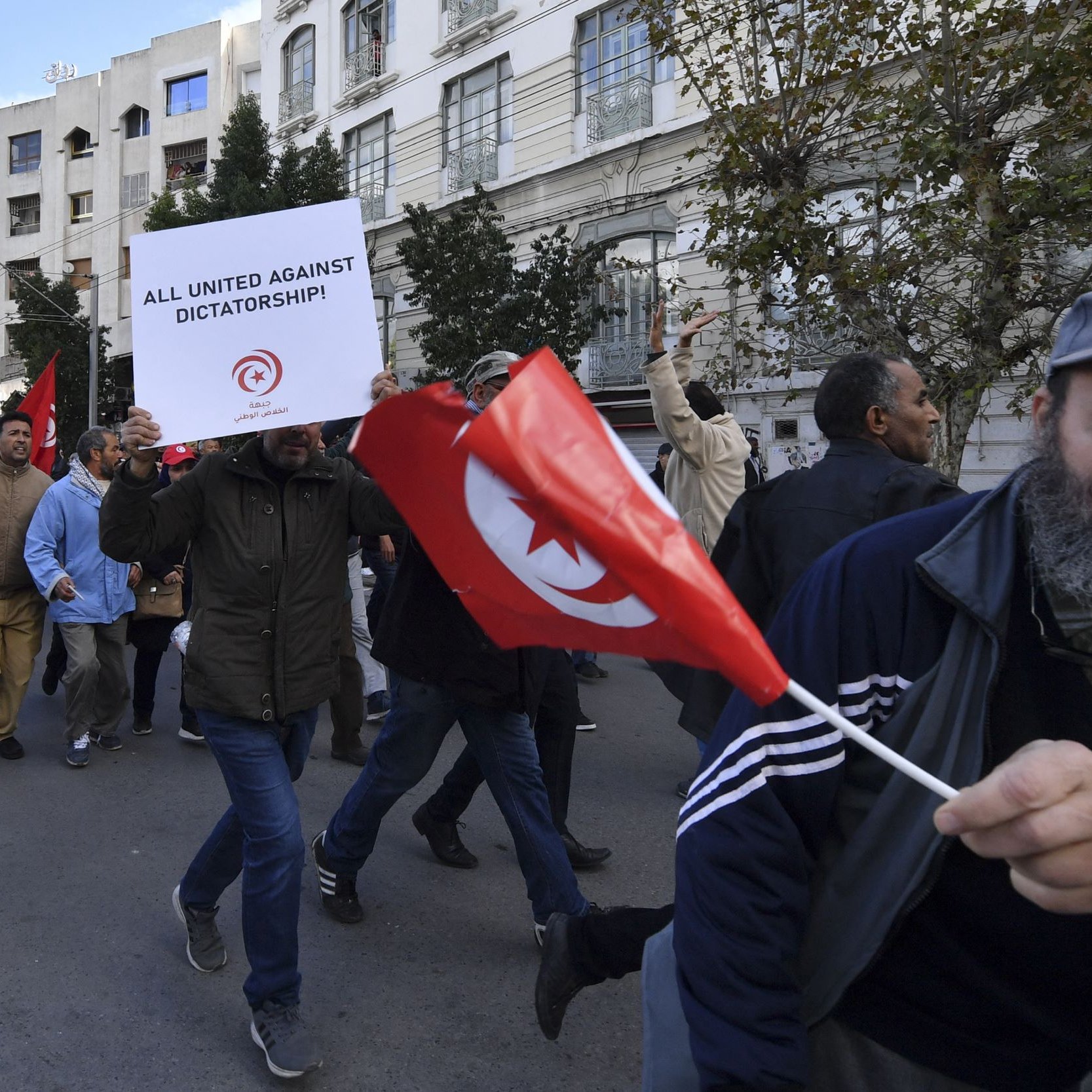 The image depicts a protest scene, likely in Tunisia. A crowd of people is marching down a street. Some participants are holding signs and flags. One prominent sign reads "ALL UNITED AGAINST DICTATORSHIP!" indicating the protest's focus on political issues. Among the crowd, a man stands out, waving a Tunisian flag and wearing a baseball cap and a jacket. The overall atmosphere suggests a movement against authoritarian rule, with participants expressing their collective dissent.