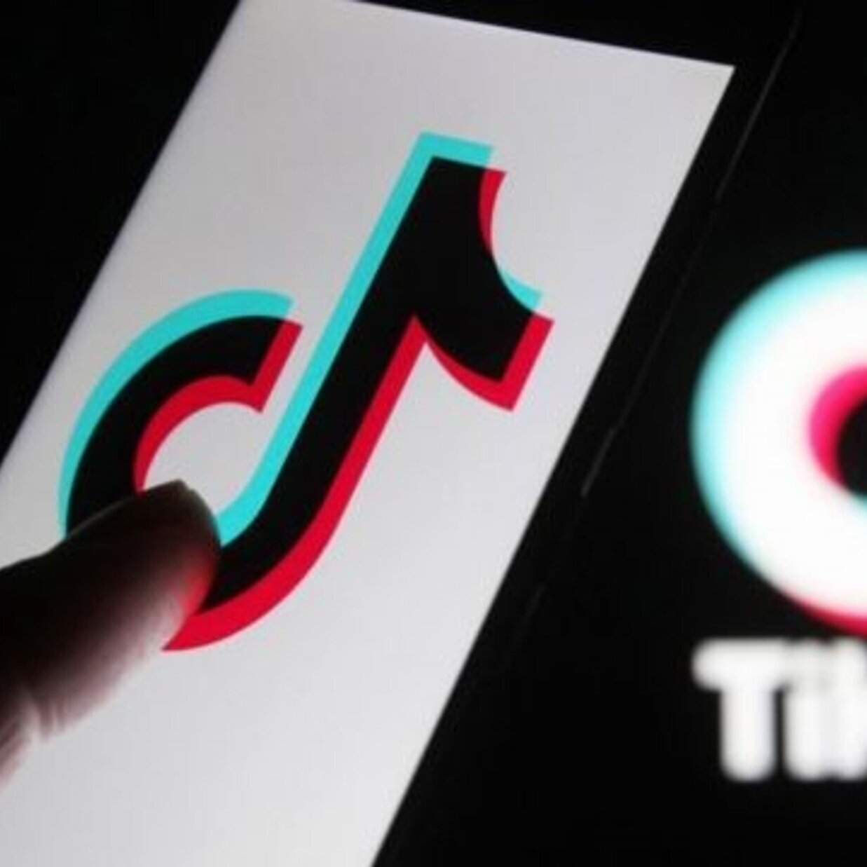 این تصویر یک گوشی هوشمند را نشان می‌دهد که لوگوی اپلیکیشن TikTok روی صفحه آن قابل مشاهده است. انگشت یک دست به سمت لوگو اشاره می‌کند. در پس‌زمینه، لوگوی TikTok به صورت بزرگ‌تر و با رنگ‌های آبی و قرمز دیده می‌شود. طراحی این لوگو به‌صورت مدرن و جذاب است و نشان‌دهنده‌ی ماهیت سرگرم‌کننده اپلیکیشن می‌باشد.