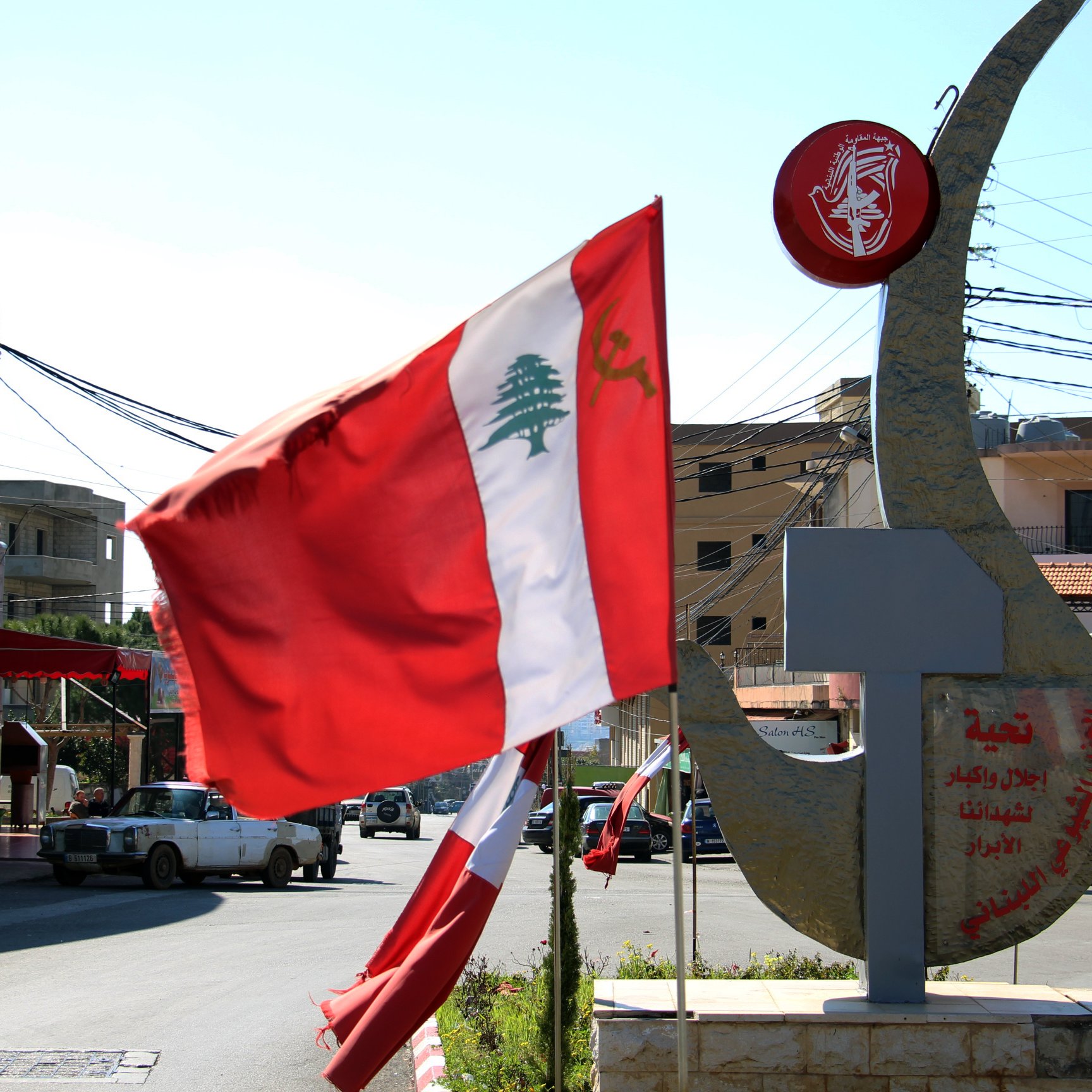 La imagen muestra una intersección en una localidad, con una bandera de Líbano ondeando en el viento. A la derecha, hay una estructura que parece una escultura o un monumento, posiblemente con inscripciones en árabe. Al fondo se pueden ver edificios y postes de luz, mientras que el cielo es despejado y soleado. La escena refleja un ambiente urbano característico de la región.