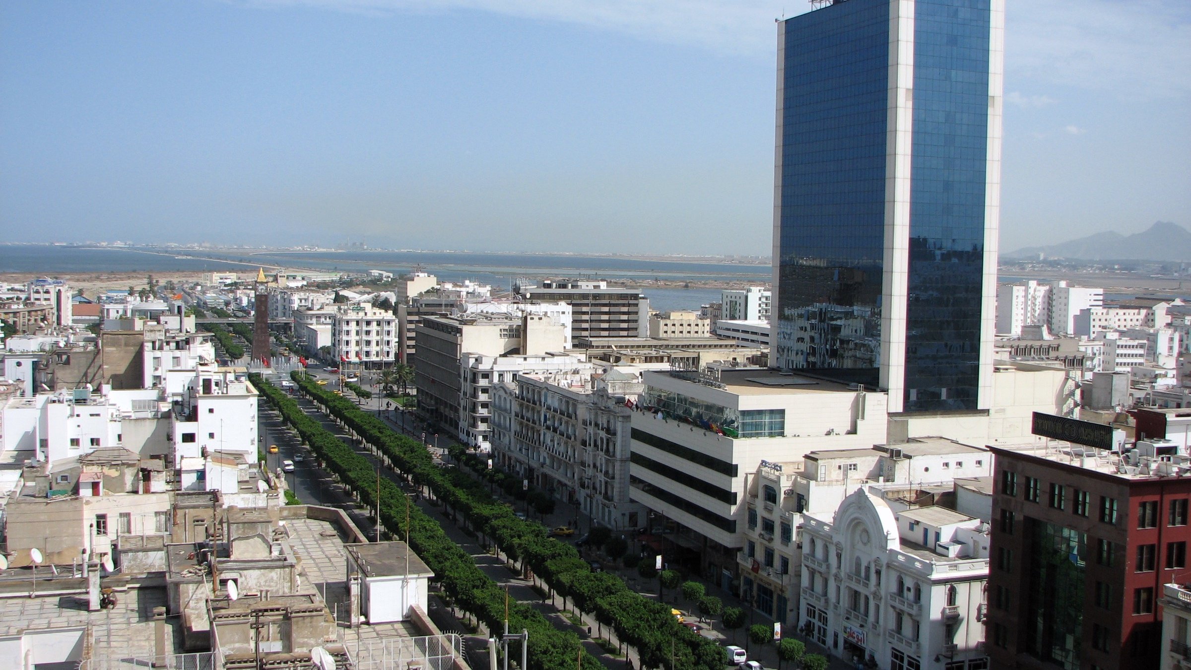 L'image montre un paysage urbain, probablement d'une ville côtière avec une vue panoramique. On peut voir de grands bâtiments modernes, dont une tour en verre qui se dresse au centre. La rue est bordée d'arbres et d'immeubles, créant une ambiance sympathique. À l'arrière-plan, on aperçoit l'océan, suggérant que la ville est située près de la mer. Le ciel est dégagé avec quelques nuages, indiquant une belle journée.